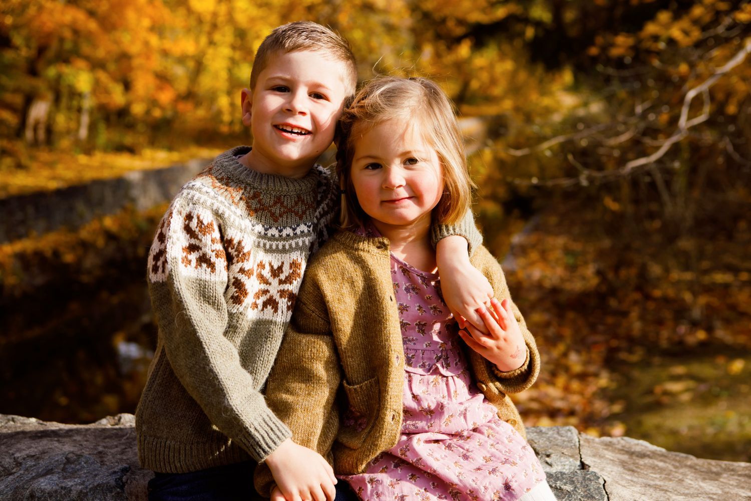 Two siblings in knit sweaters sitting together on a log with colorful autumn trees behind them.