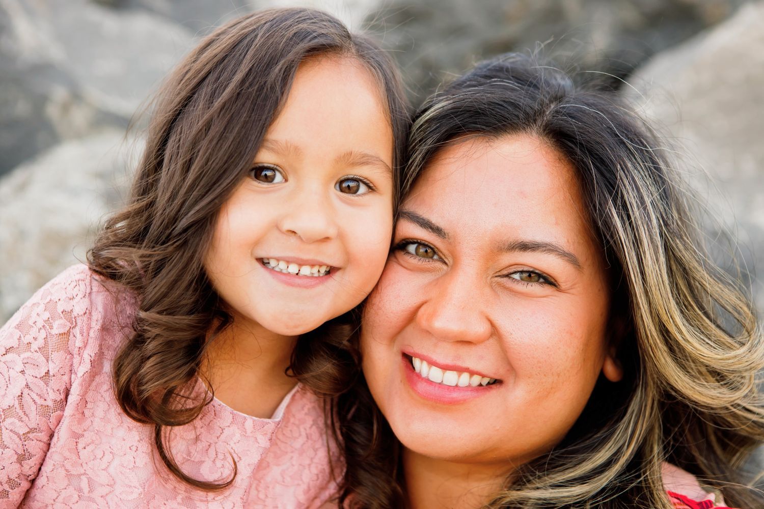 Smiling Hispanic mother and young daughter in pink pose cheek to cheek outdoors.