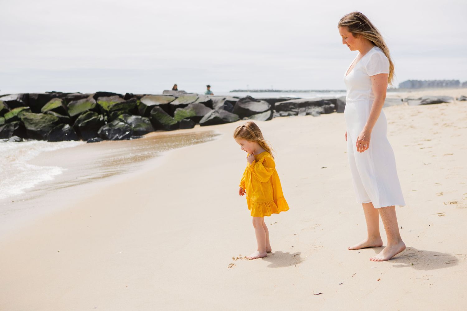 A pregnant woman in a white dress watching her young daughter in yellow on a sandy beach.