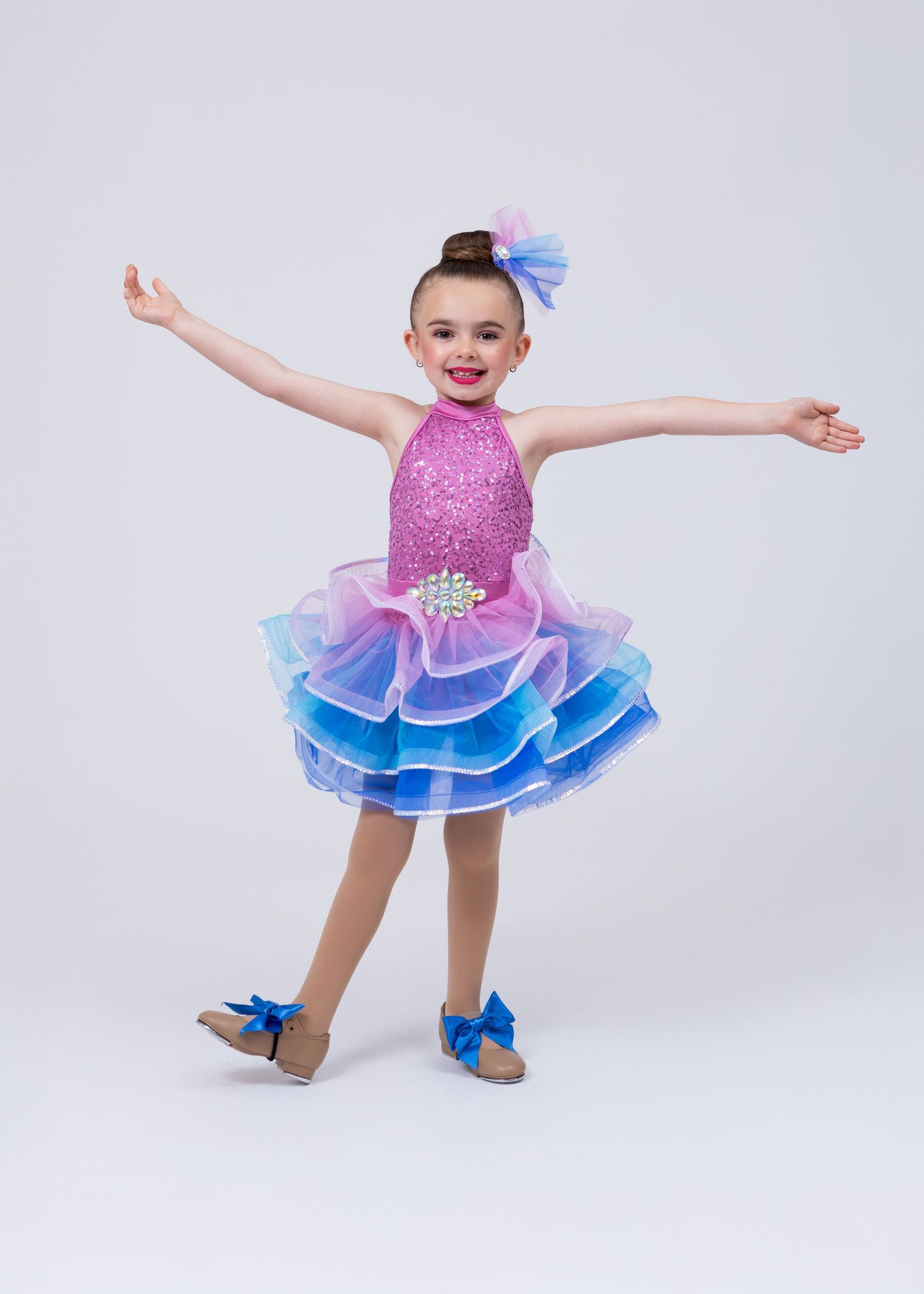 A young girl in a pink sequined tiered tutu dress poses with arms outstretched on a white background.