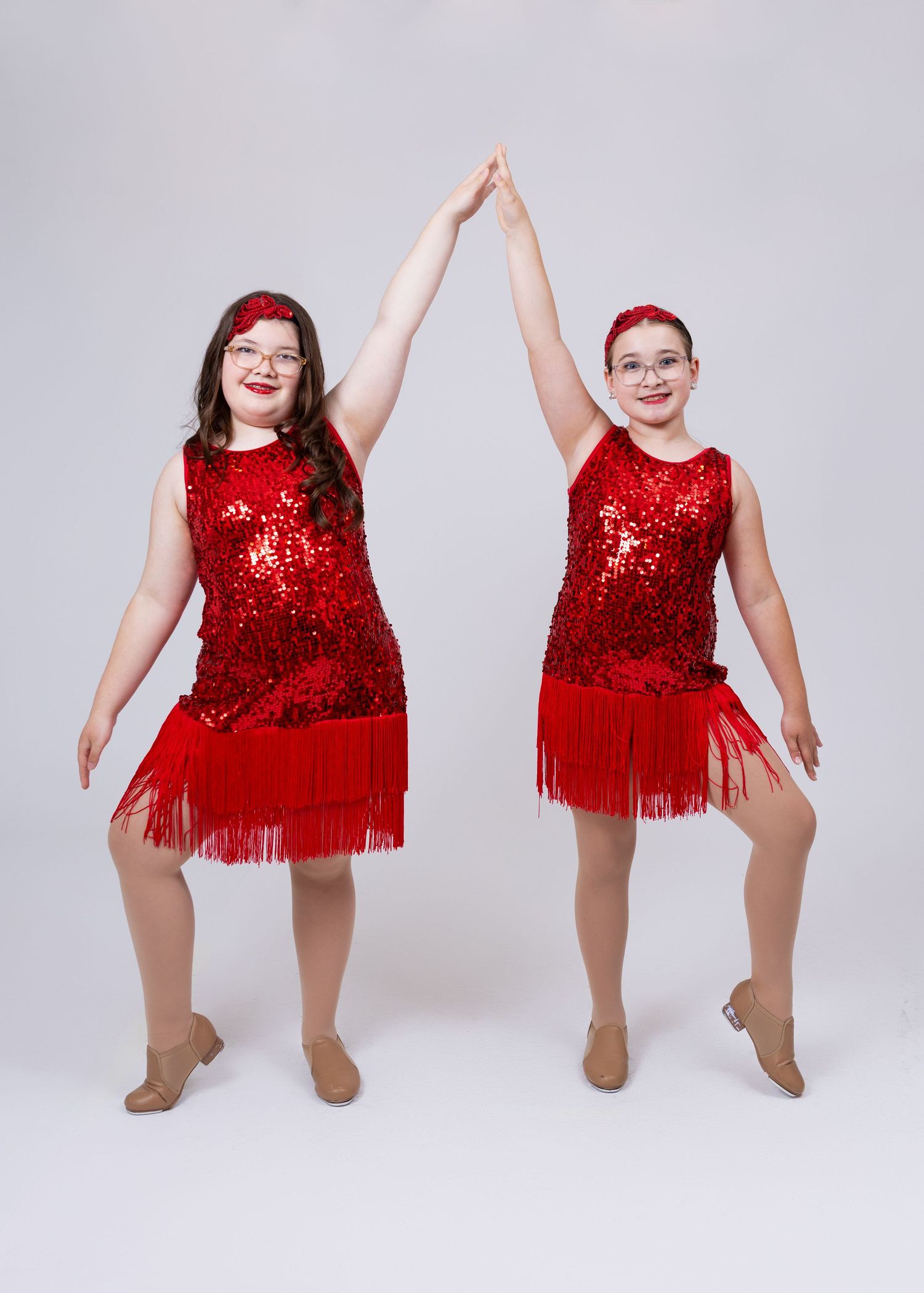 Two girls in matching red sequin fringe dresses hold hands raised in a dance studio photo.