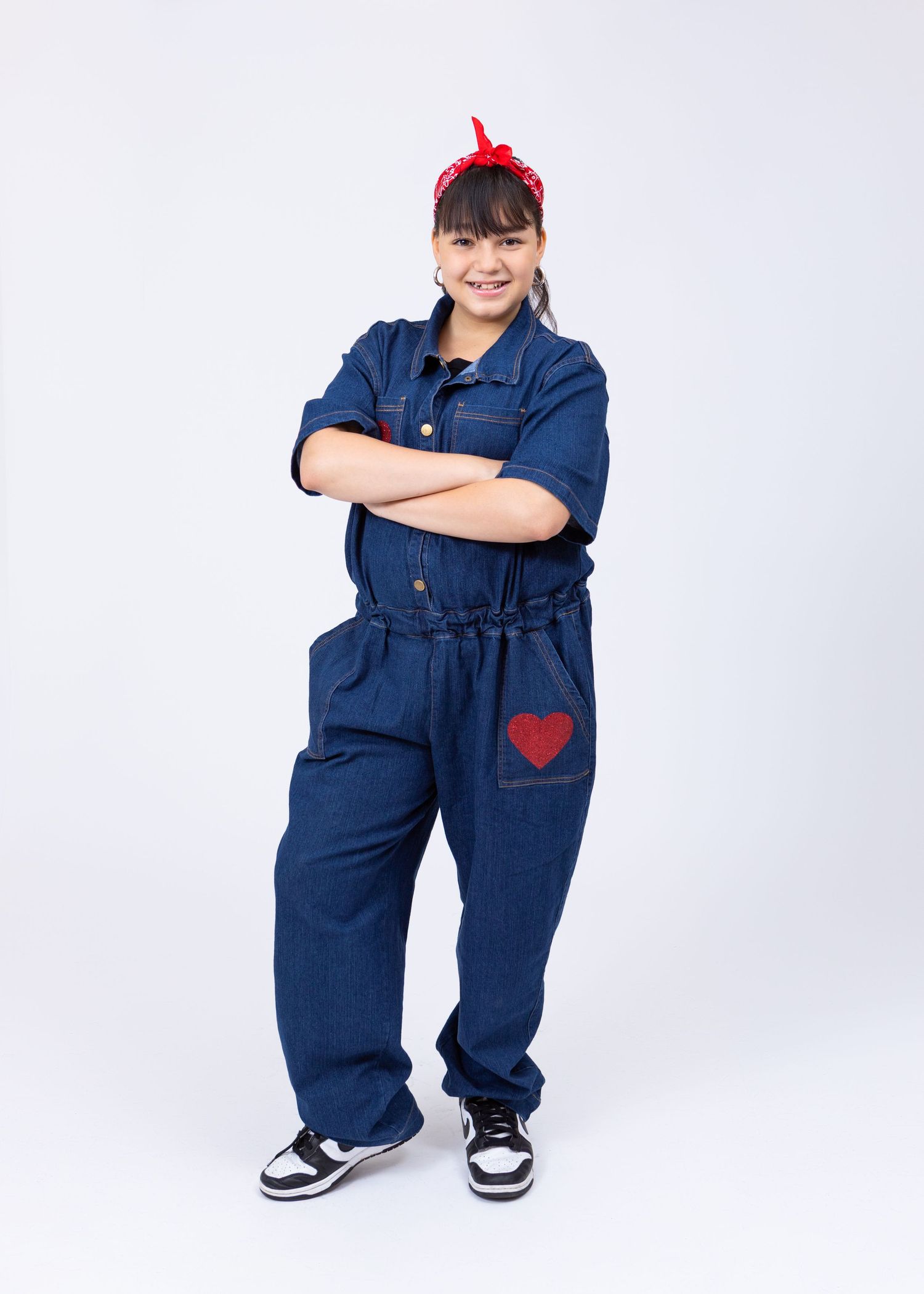Confident girl in denim jumpsuit with red heart patch and bandana poses with arms crossed.