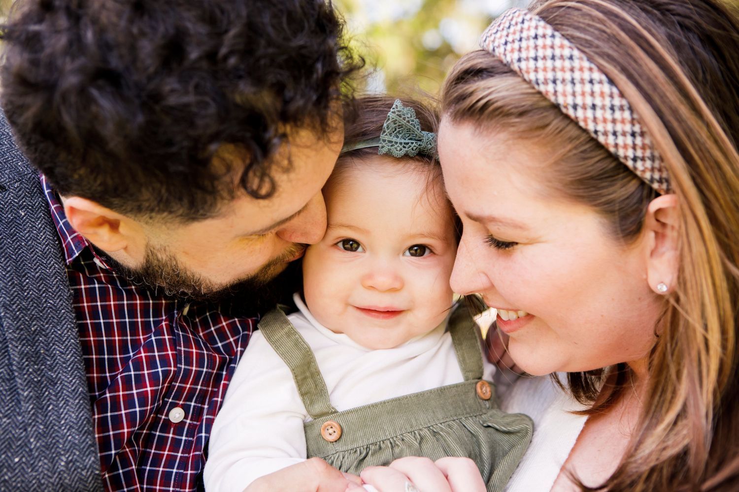 Parents lovingly kiss their smiling baby girl on cheeks in warm outdoor family portrait.