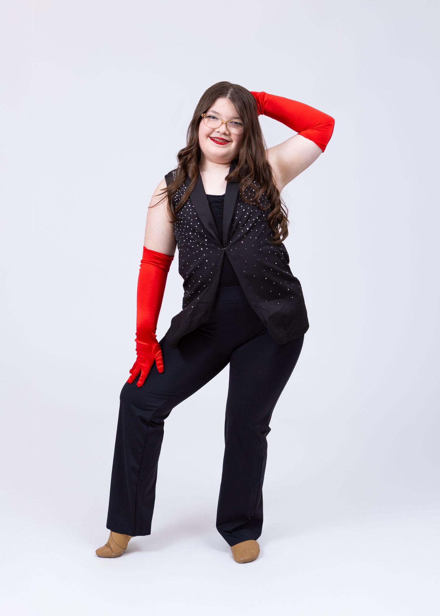 A dancer poses confidently in black sequin vest, pants, and long red gloves in a studio.
