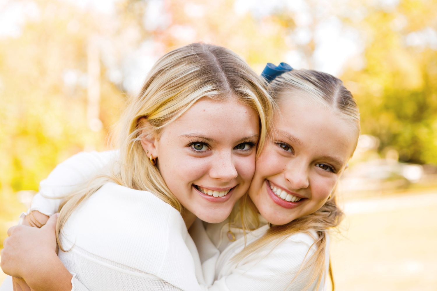 Two blonde girls hugging and smiling warmly outdoors with bright yellow autumn trees.