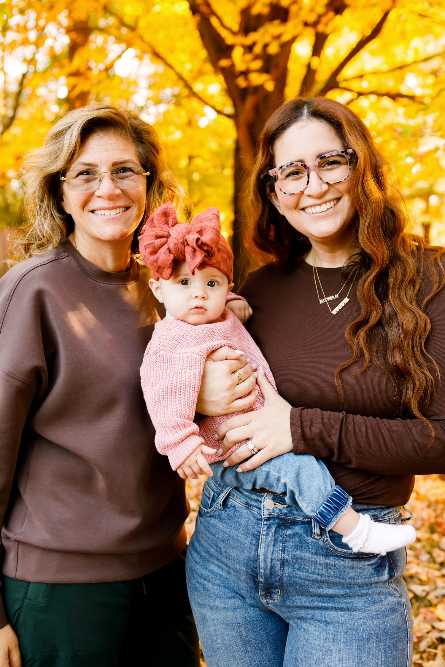 Two women and baby girl posing before vibrant yellow fall foliage.