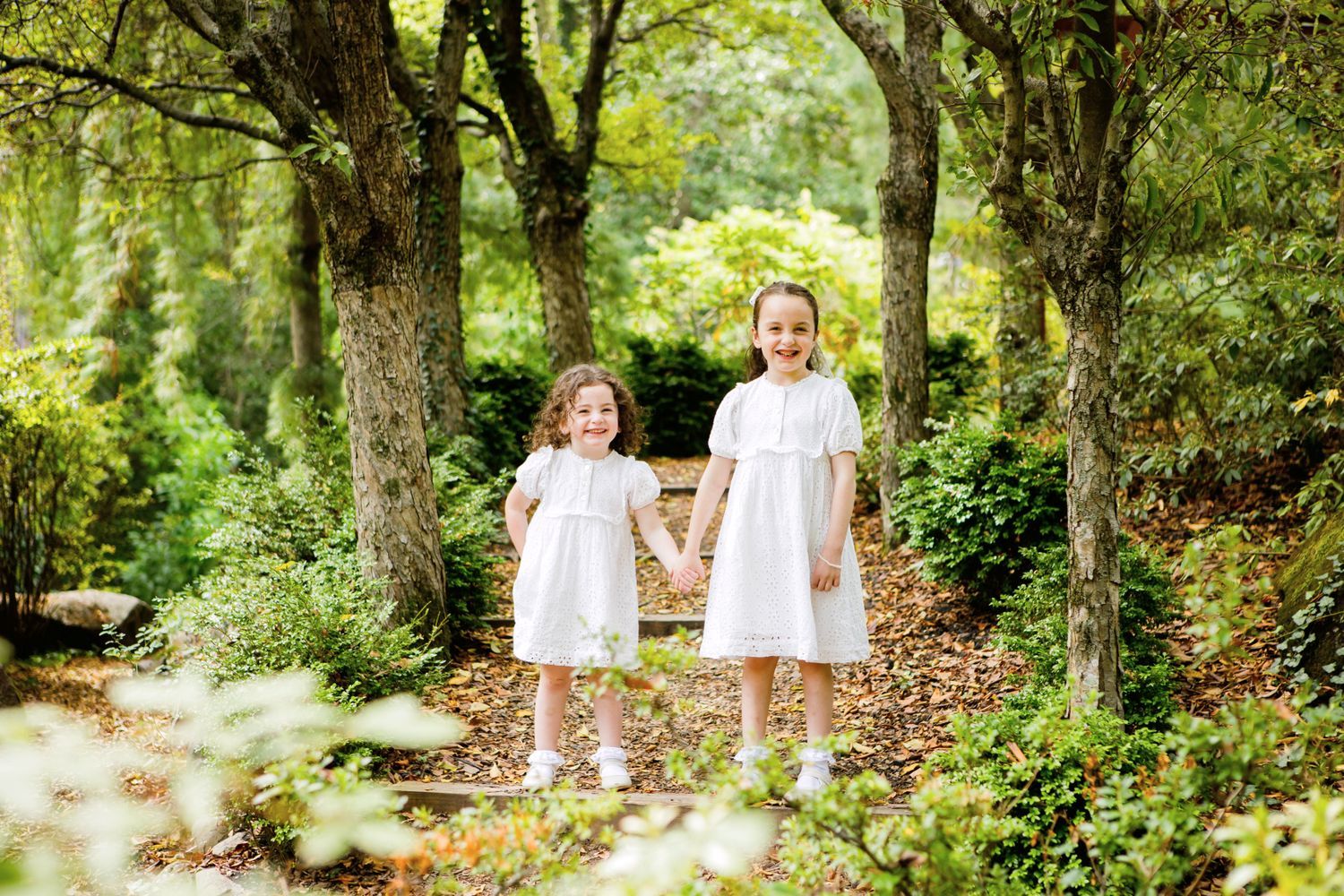 Two young girls in white dresses holding hands in a lush green garden.