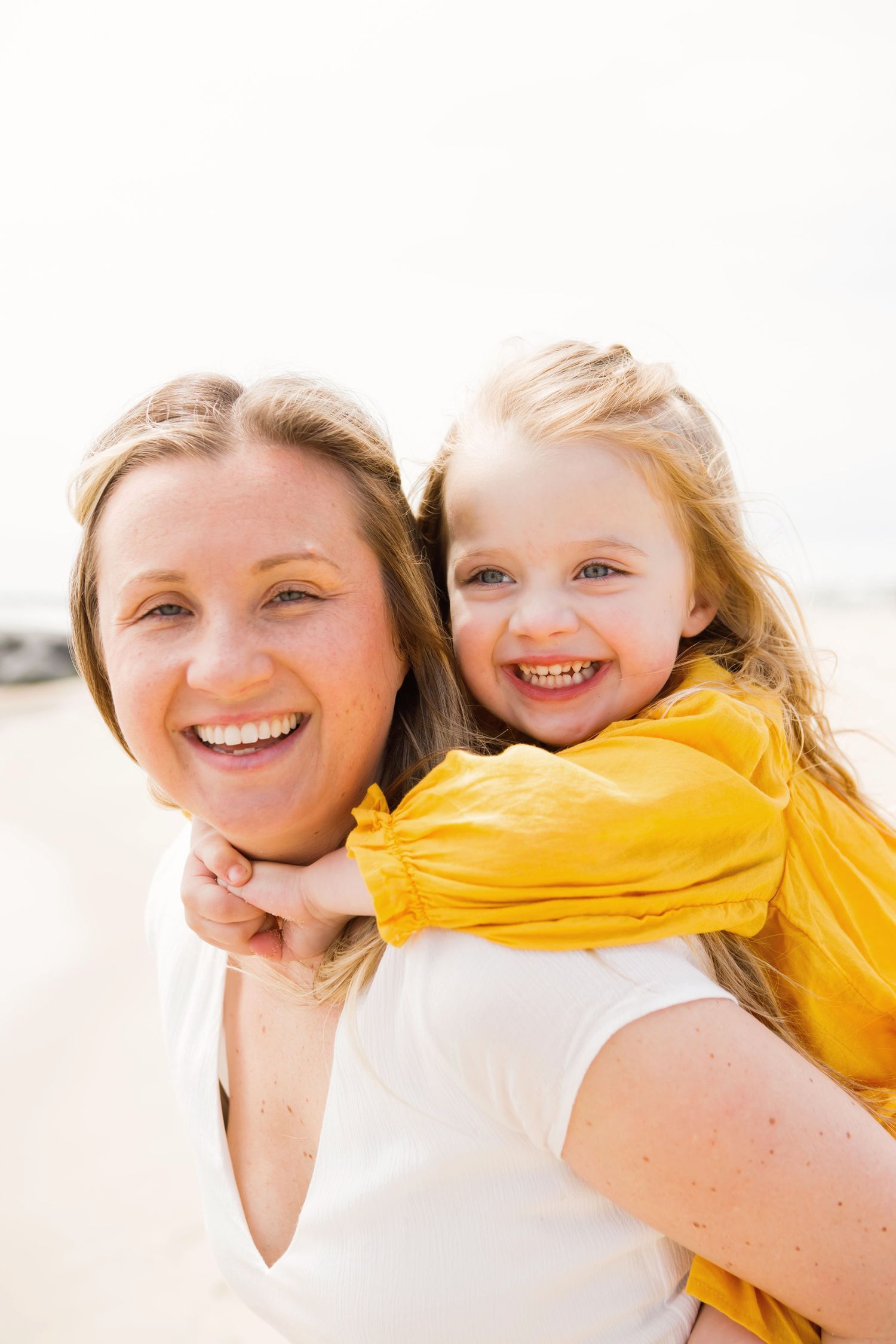 A smiling mother giving her laughing daughter a piggyback ride on a bright sunny beach.