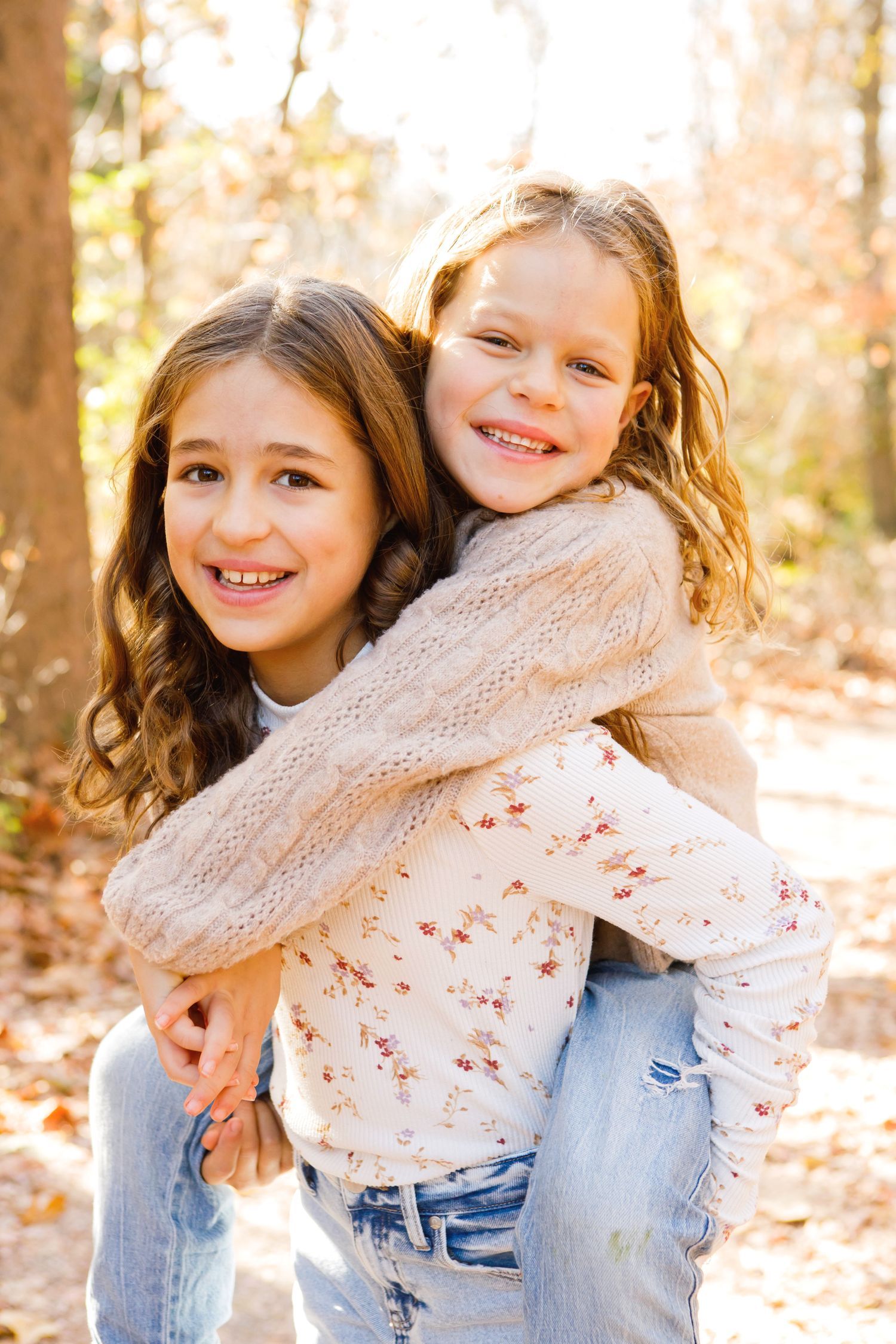 Two smiling sisters posing together with a piggyback ride in a sunny autumn forest.
