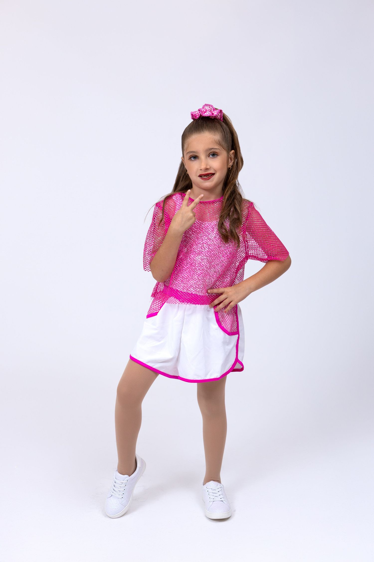 Young girl in pink sequin top and white skirt dance costume points finger playfully.