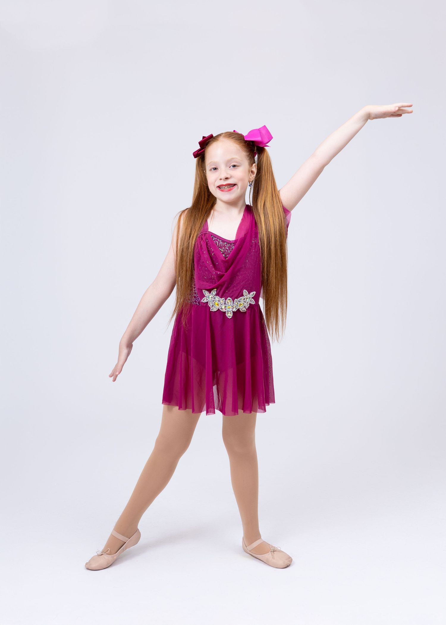 Young red-haired girl in magenta lyrical dance dress extends arms in elegant ballet pose.
