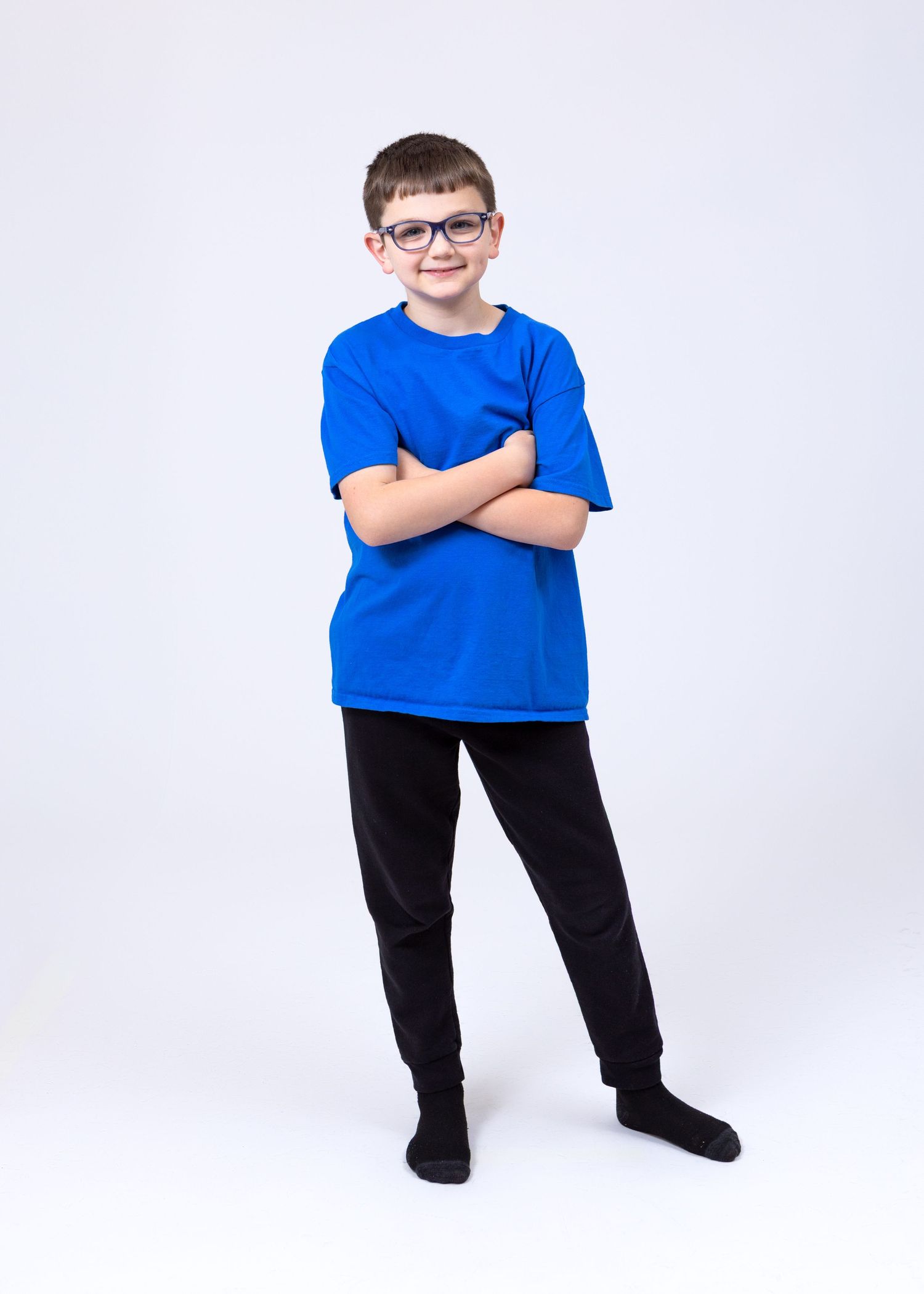 A young boy wearing glasses, a blue t-shirt, and black pants stands with arms crossed smiling.