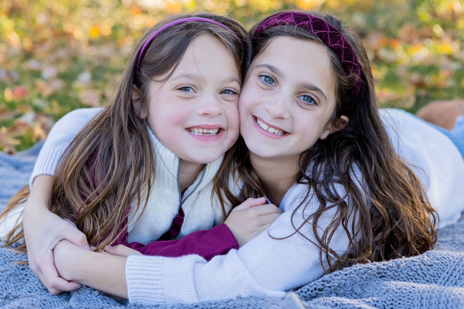 Two young sisters with brown hair smile and hug while lying on blanket in autumn park.