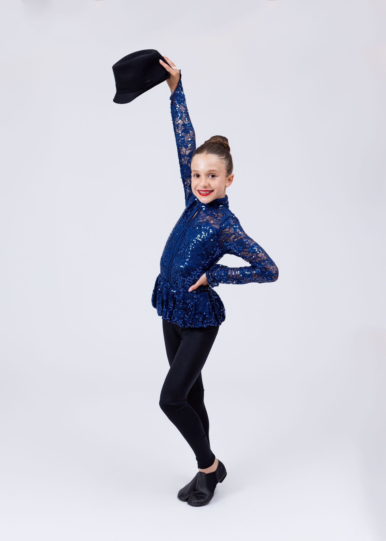 Young girl in blue sequin lace top and black leggings holds a hat up in a dance pose.