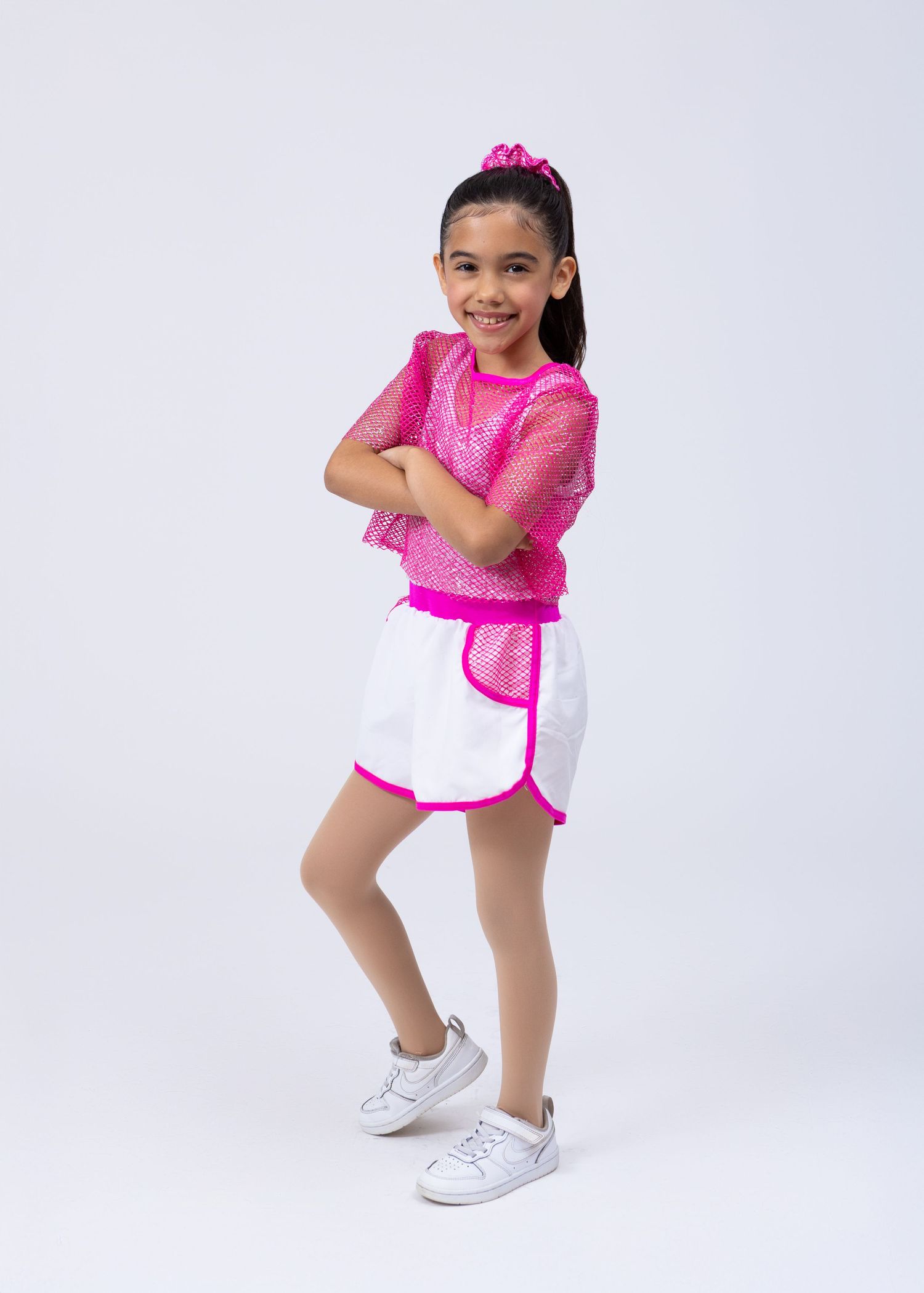 A young girl in a pink mesh top and white shorts with pink trim poses with arms crossed, smiling.