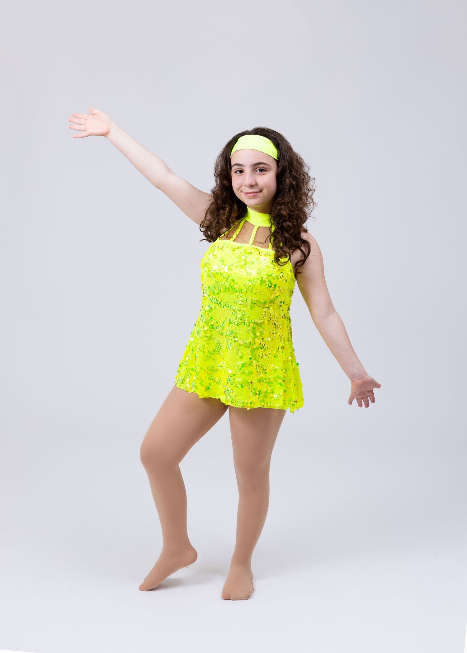 A girl in a neon yellow sequined dance costume and headband reaches one arm out while posing.