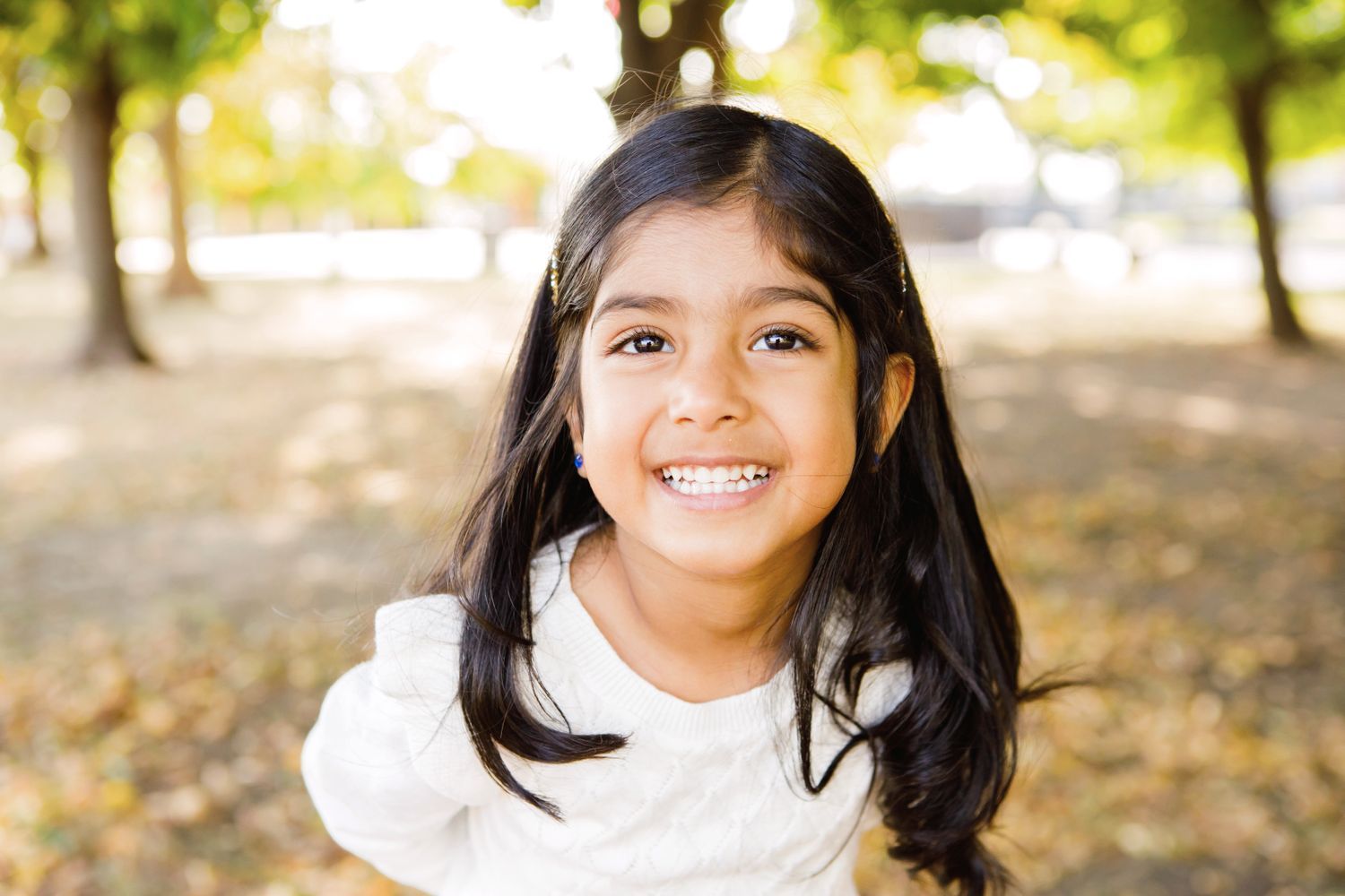 A cheerful young girl with long dark hair smiling in a sunny autumn park setting.