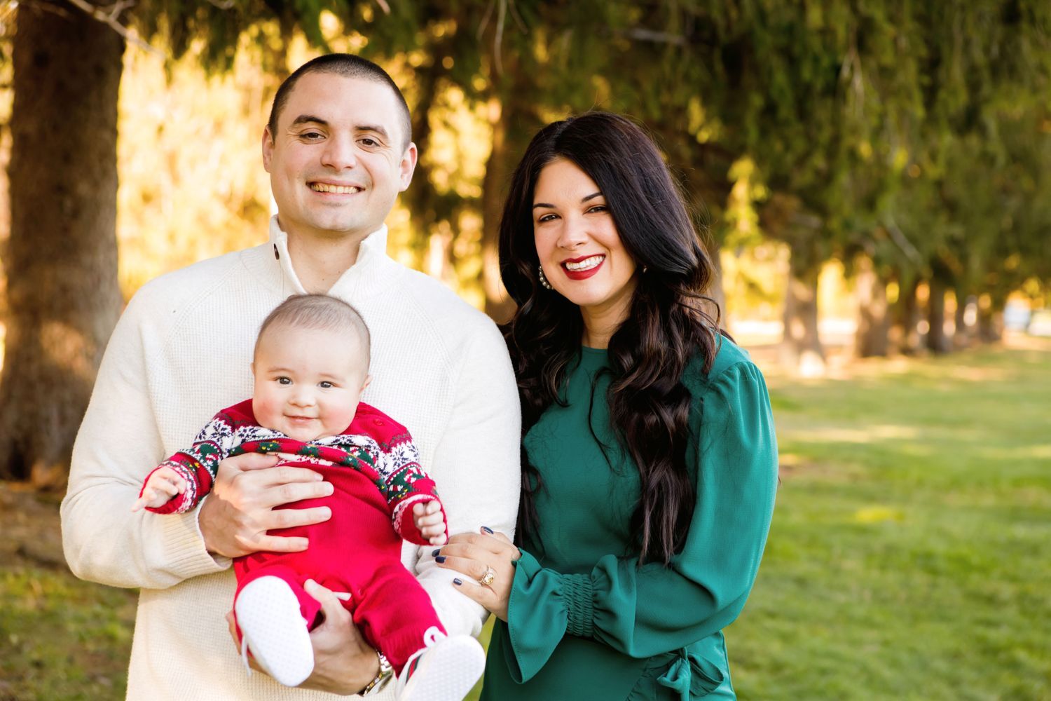 Couple smiles holding infant in red outfit during outdoor fall family portrait among tall trees.