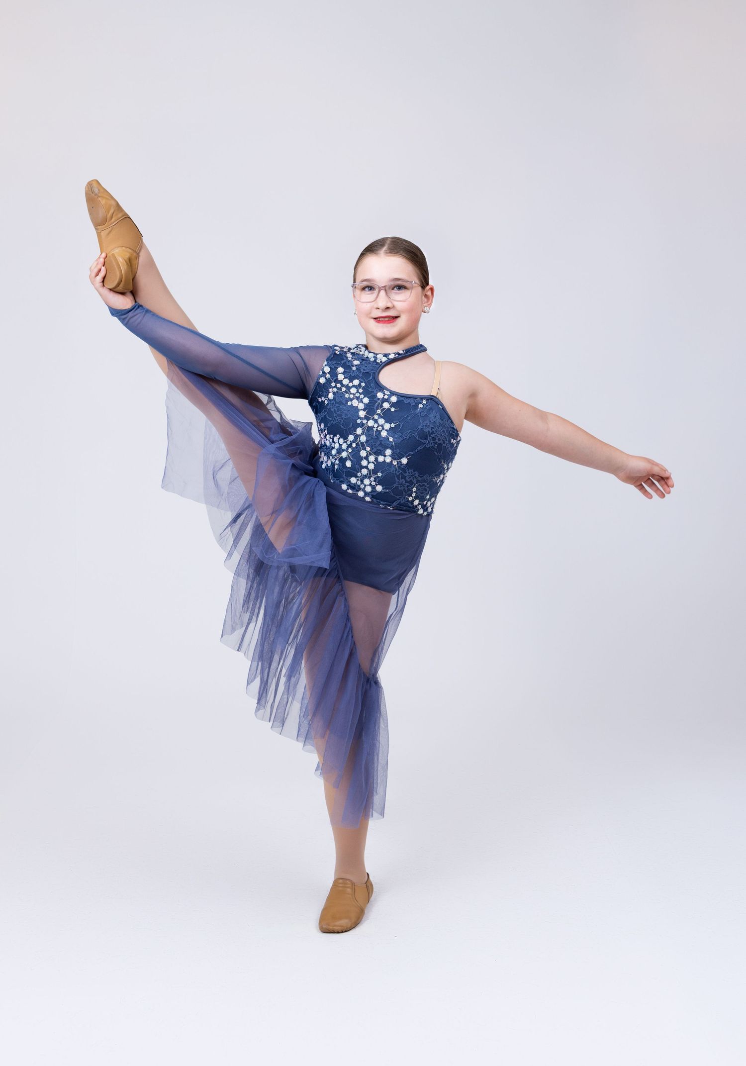 A dancer in a blue floral lyrical costume performs a high leg extension in tan shoes.