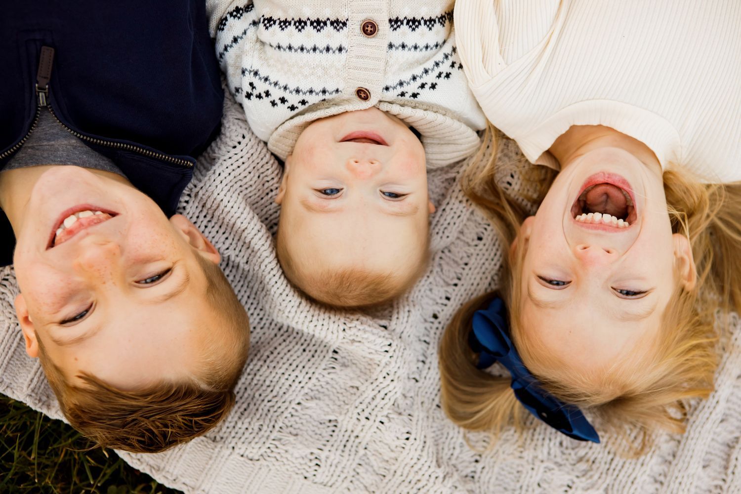 Three young children laugh and smile lying upside down together on grass in outdoor portrait.