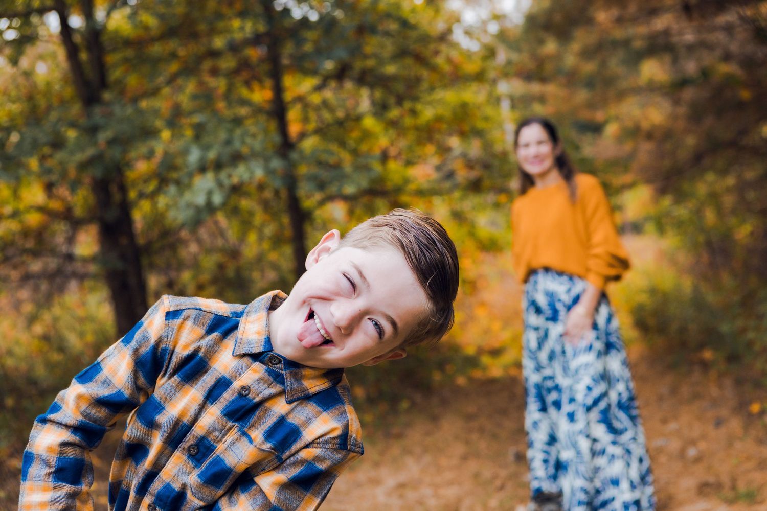 A playful boy in a plaid shirt makes a funny face outdoors while a woman in orange stands in the background.