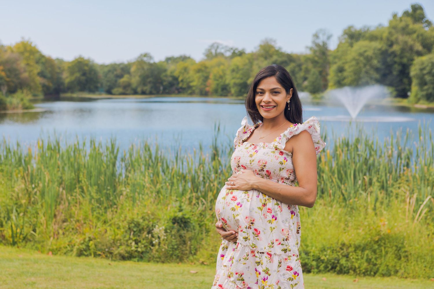 A smiling pregnant woman in a floral dress cradles her baby bump near a tranquil lake outdoors.