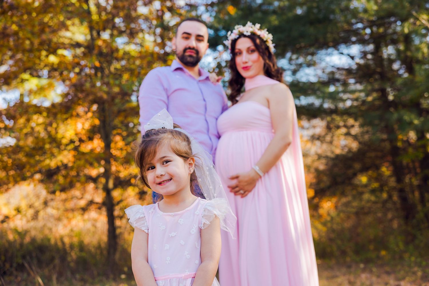 Pregnant woman in pink gown with flower crown posing with husband and daughter outdoors.