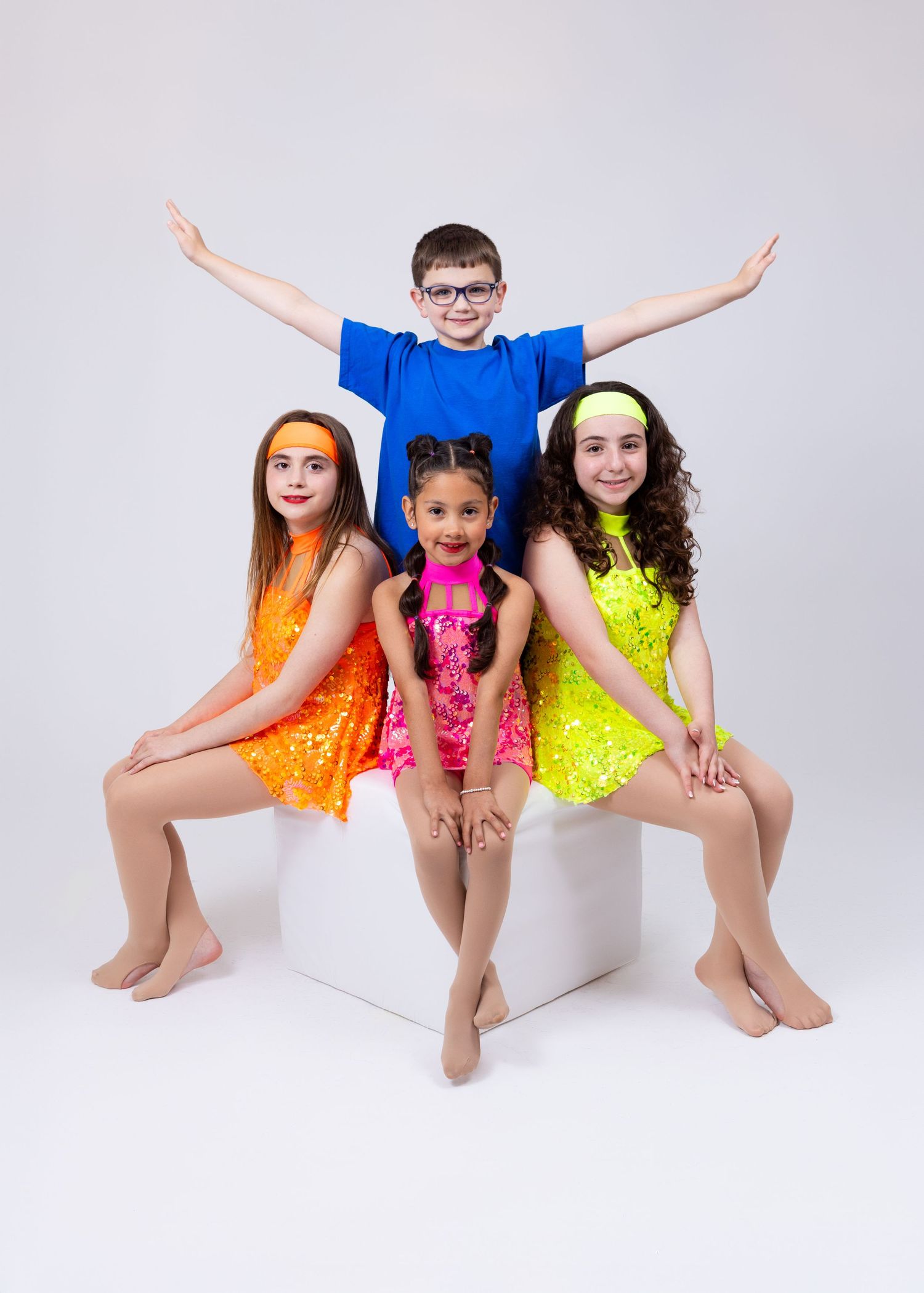 Four young dancers in colorful sequin costumes pose together on a white cube for a studio photo.