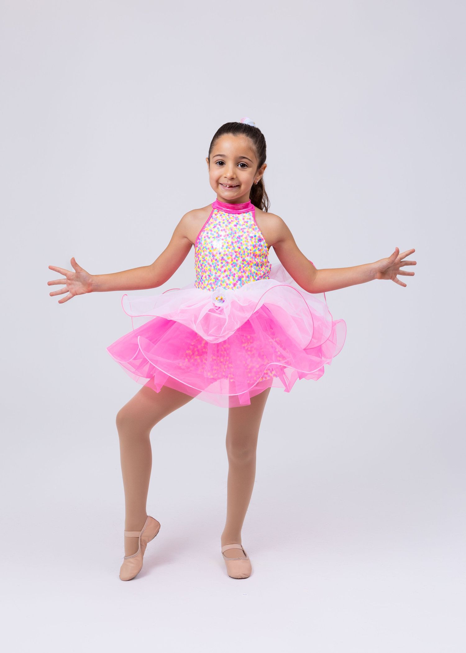 Young girl in pink sequined halter dance costume performing ballet pose on white background.