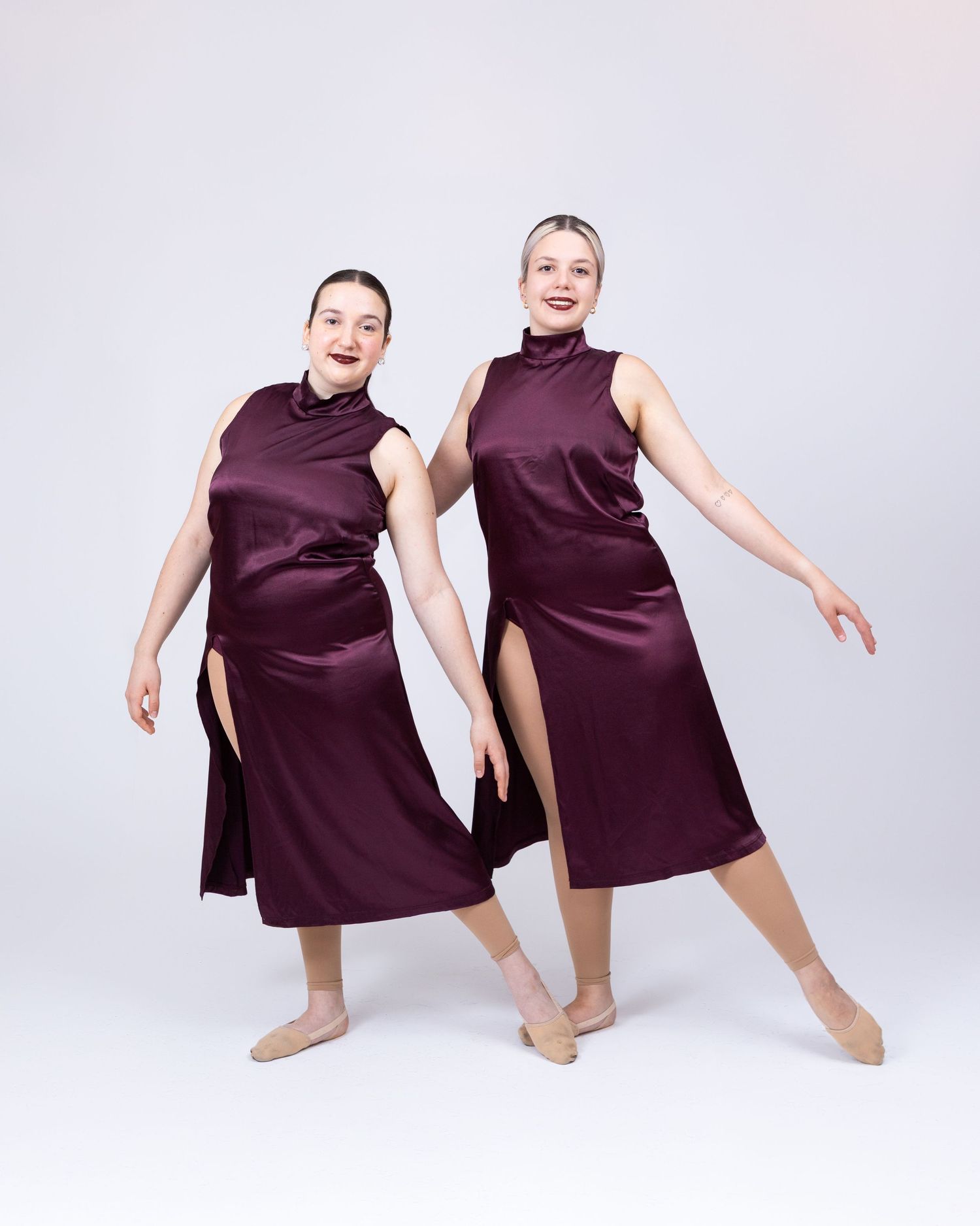Two female dancers in matching burgundy satin midi dresses pose gracefully against a white studio background.