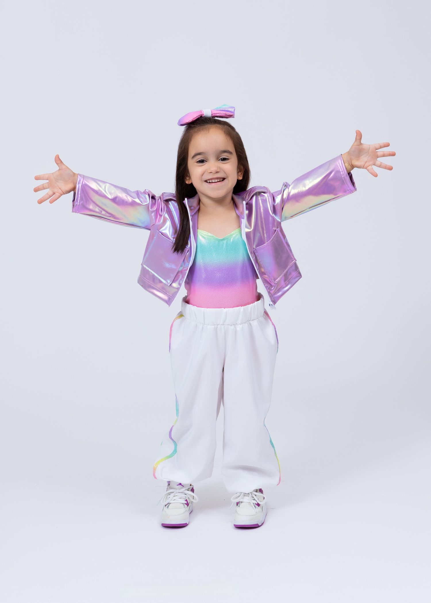 Happy girl in iridescent purple jacket, colorful crop top, and white pants poses with arms wide open.