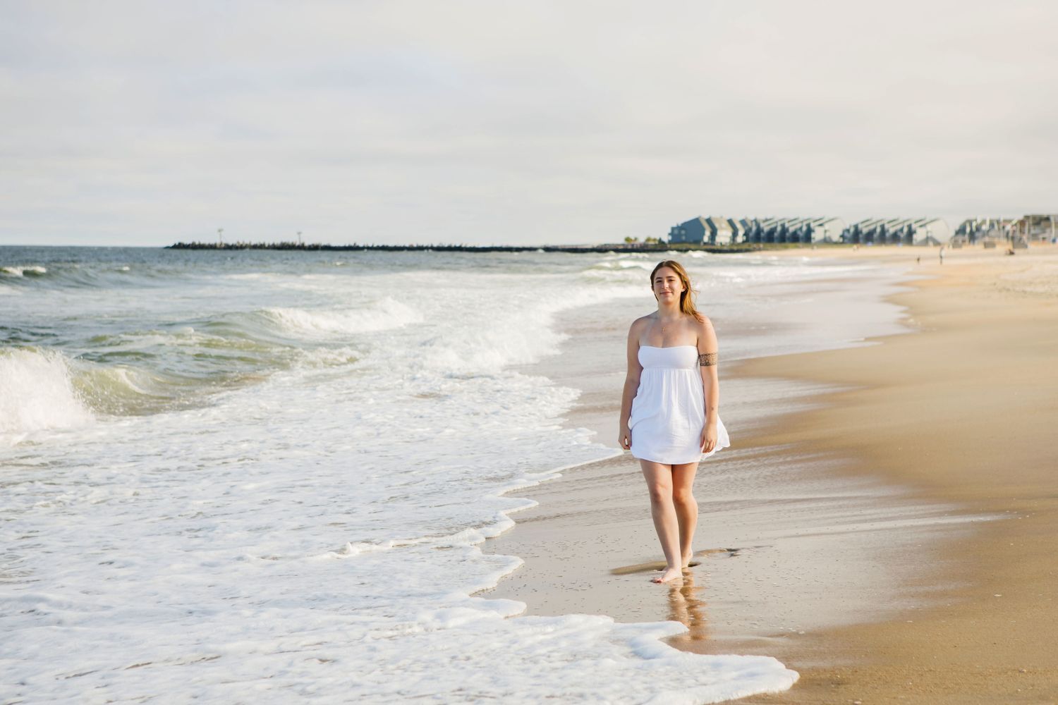 A young woman in a white dress walking barefoot along a scenic sandy ocean beach.