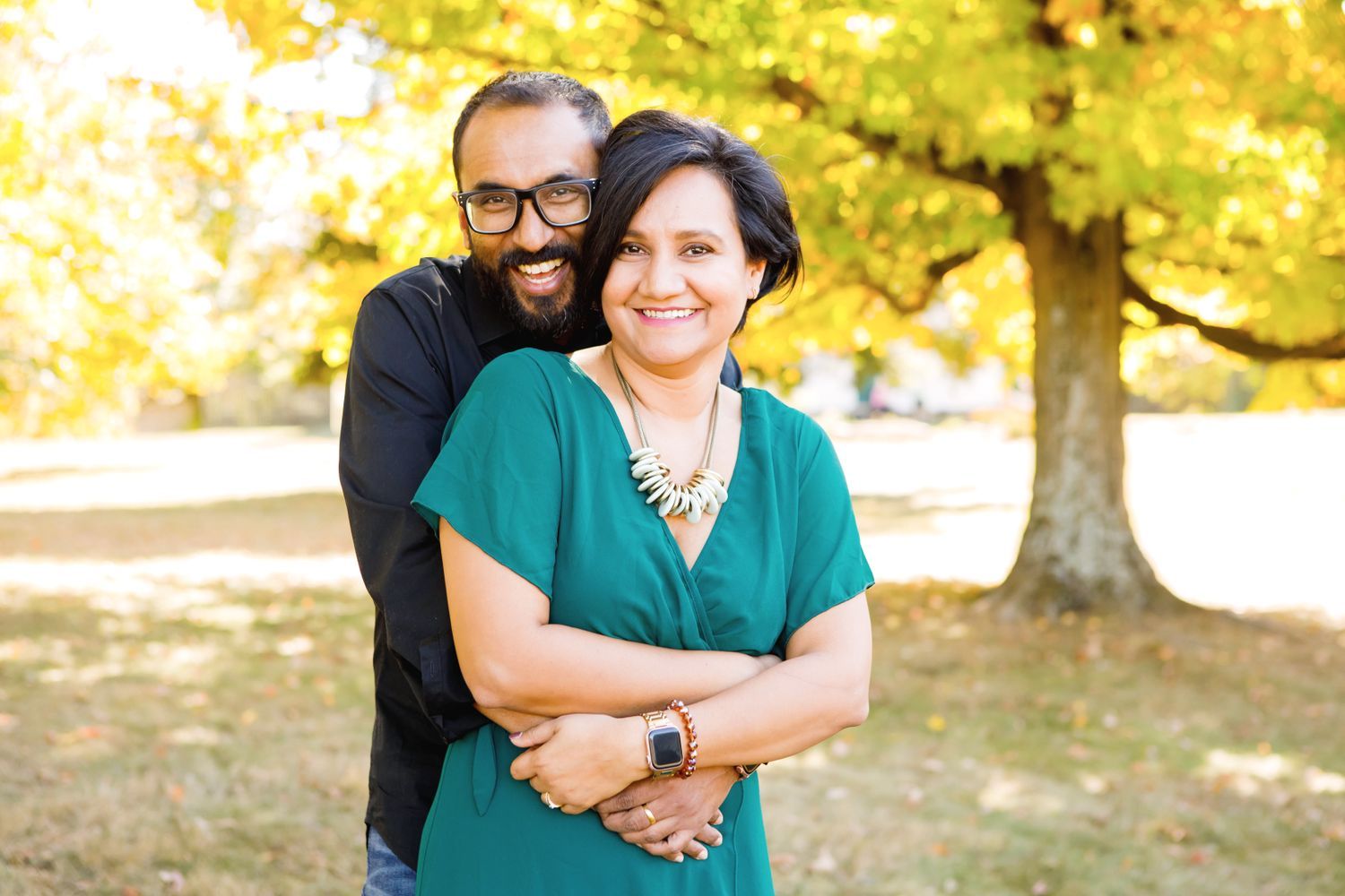Smiling couple embracing outdoors under a large tree with golden fall foliage.