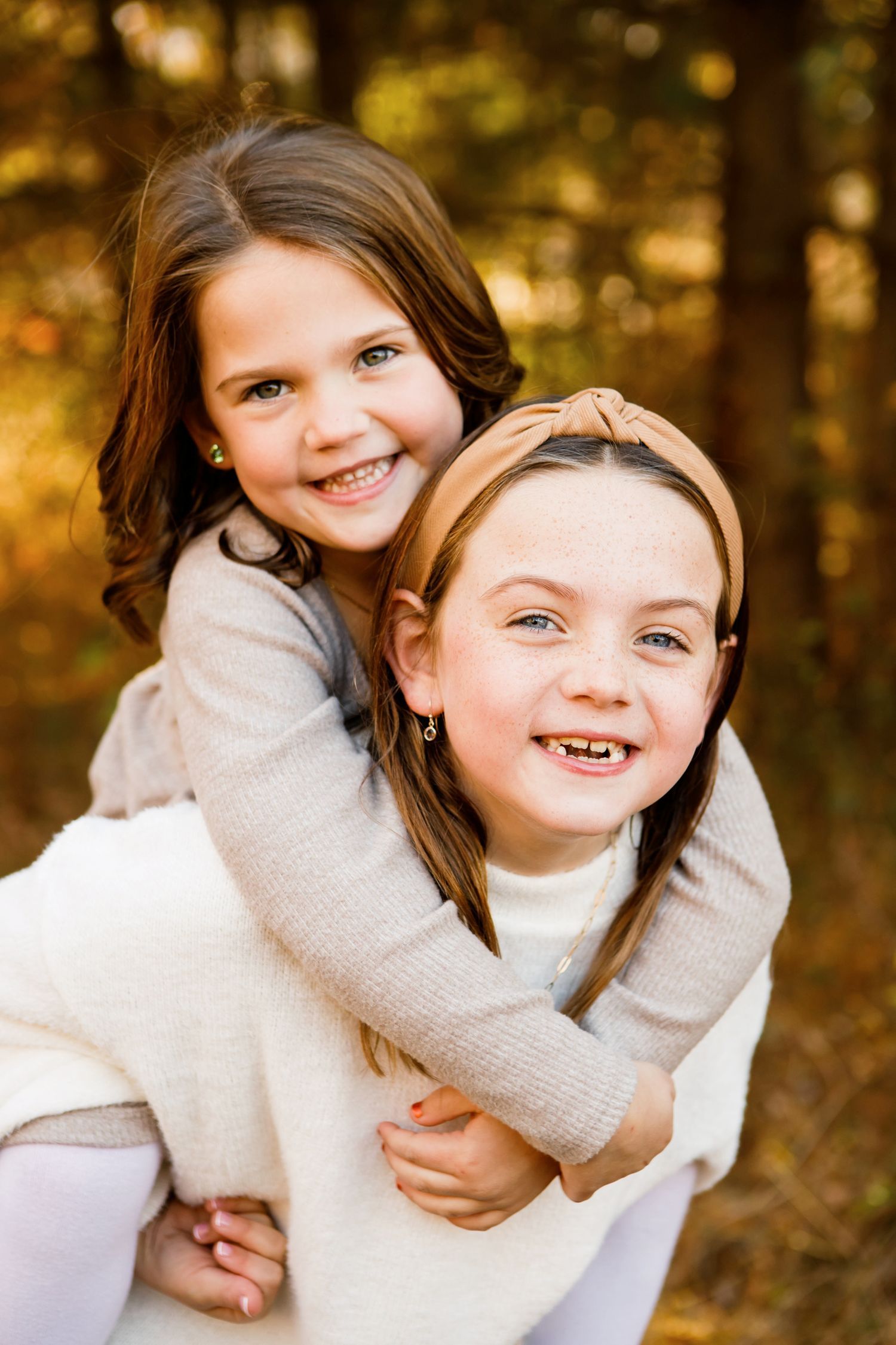 Two young sisters smiling, one giving the other a piggyback ride in an autumn forest.