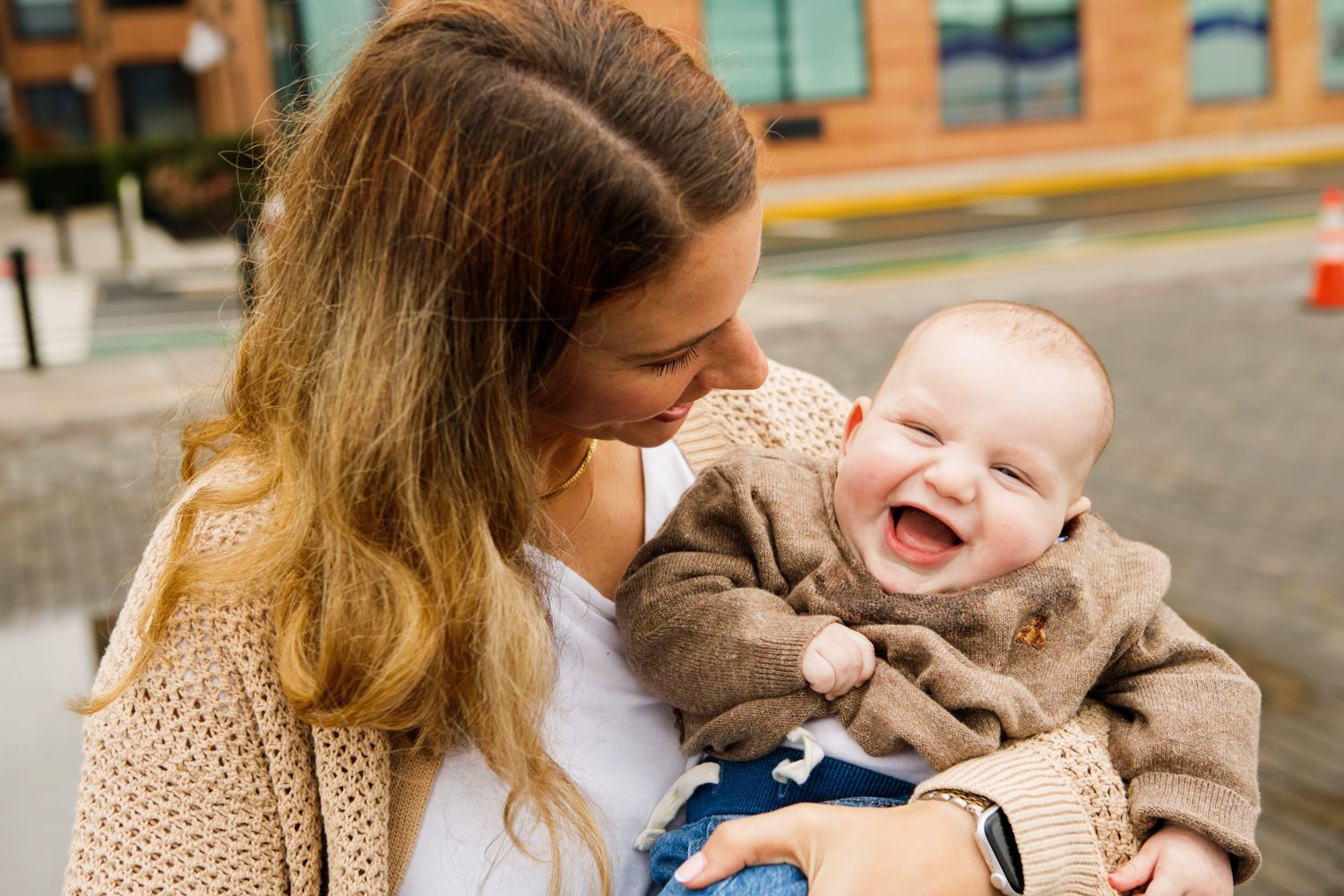 Joyful baby in brown jacket laughs while being held by smiling mother outdoors.