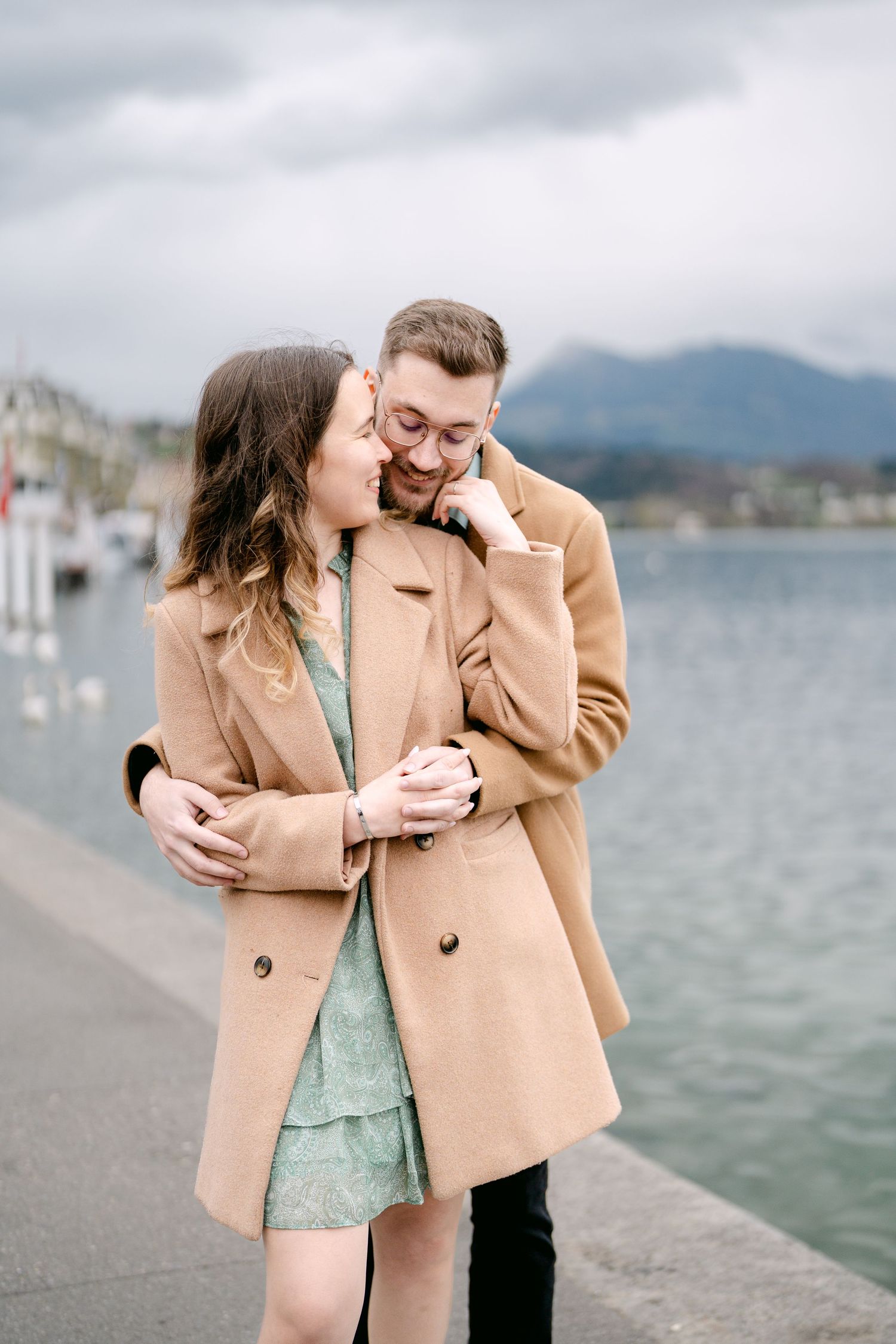 A couple in camel coats shares a laugh by the waterfront with mountains and boats in background.