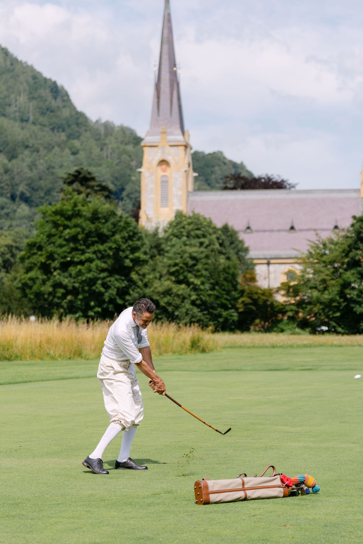 Player using a mallet during a croquet match on a sunny day with church spires in the background.