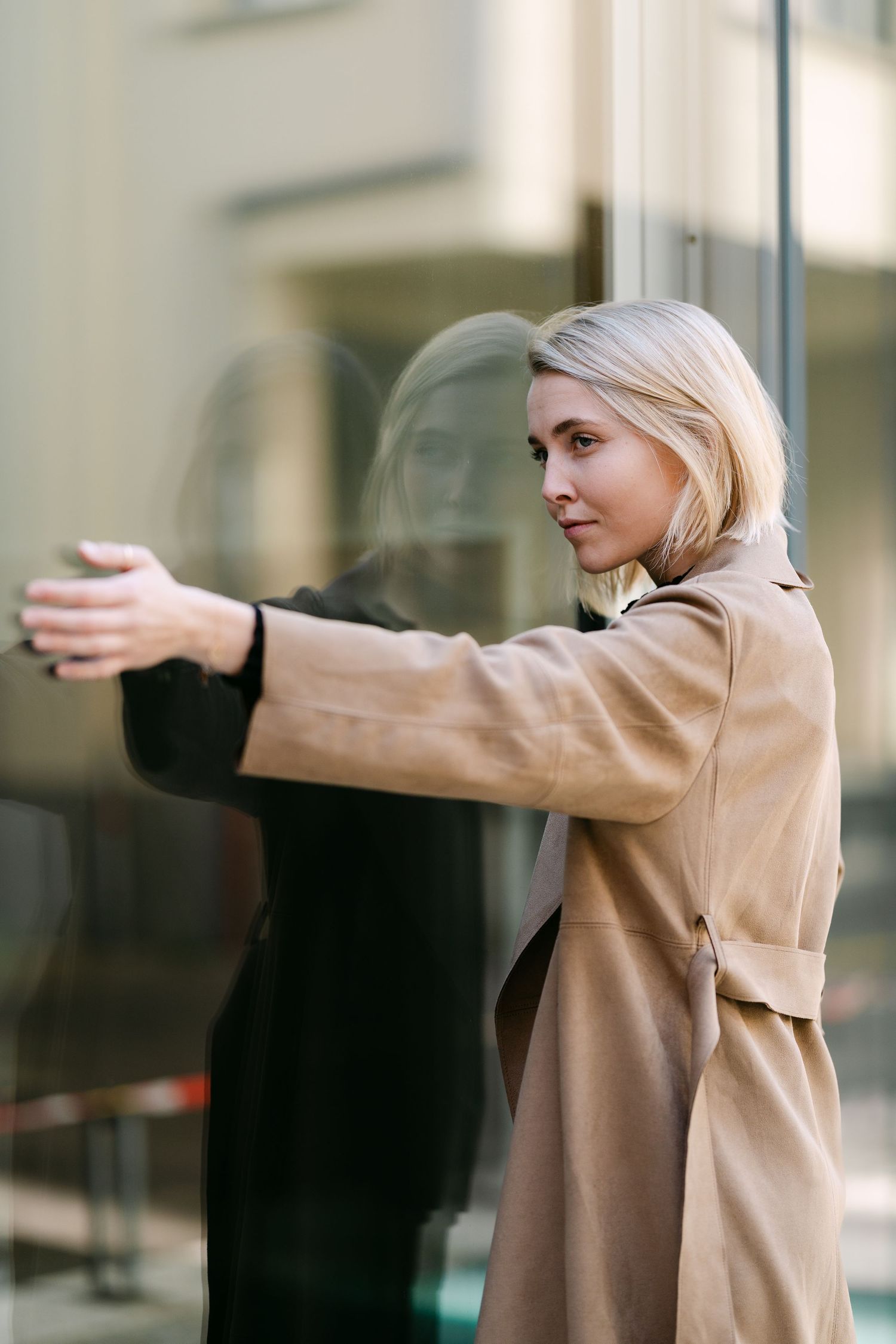Person in beige coat stretches arms outward against a light-colored building wall.