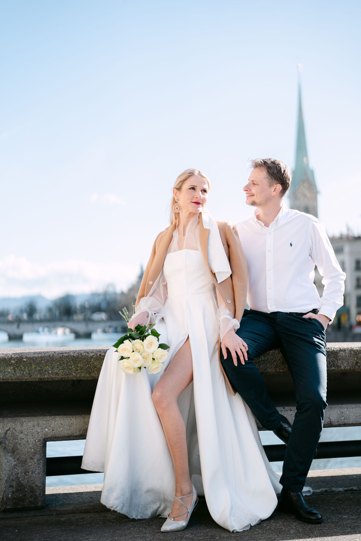 Elegant wedding couple sitting on stone wall with city skyline and church spire in background.