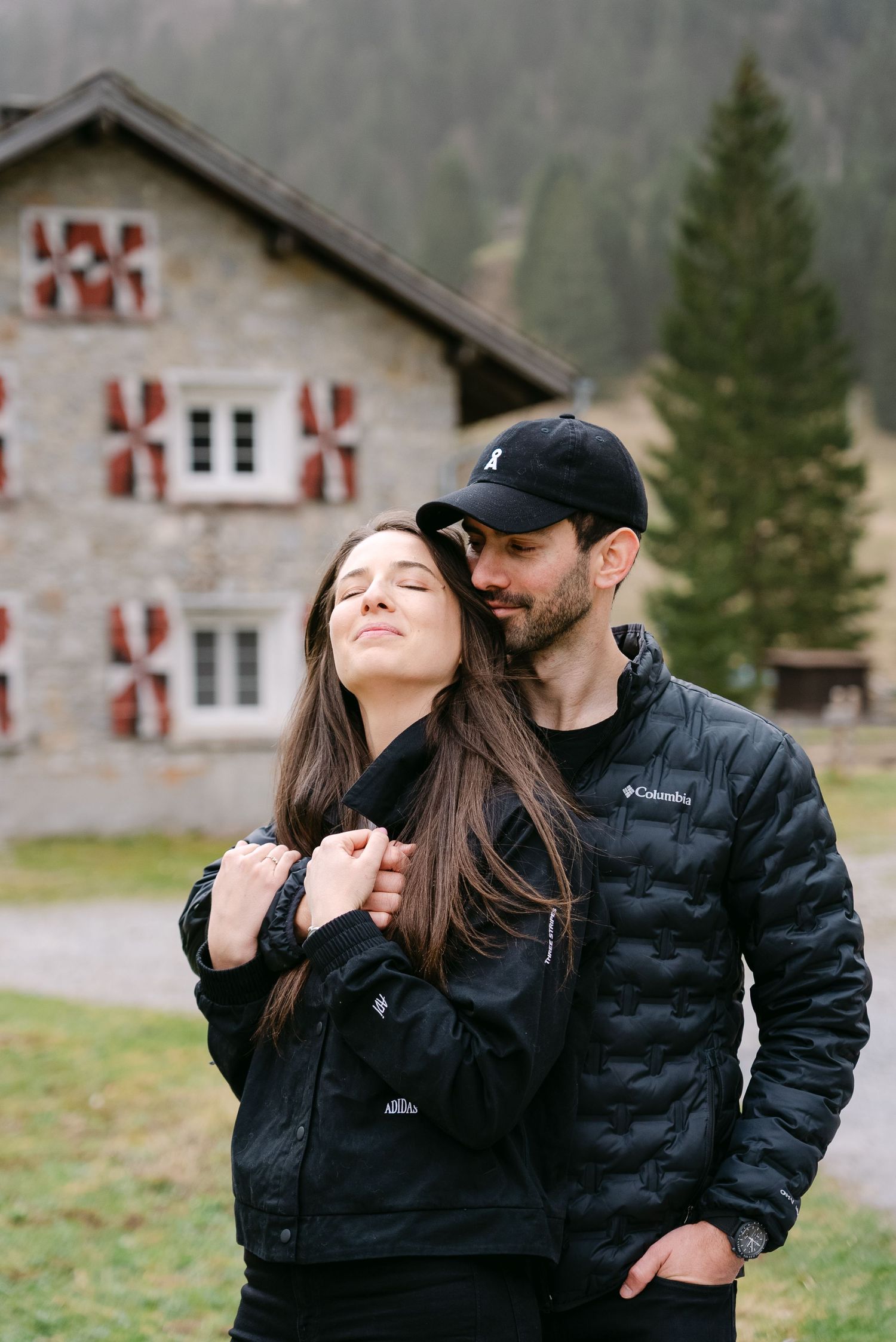 Couple in dark winter jackets embracing near a rustic mountain building.