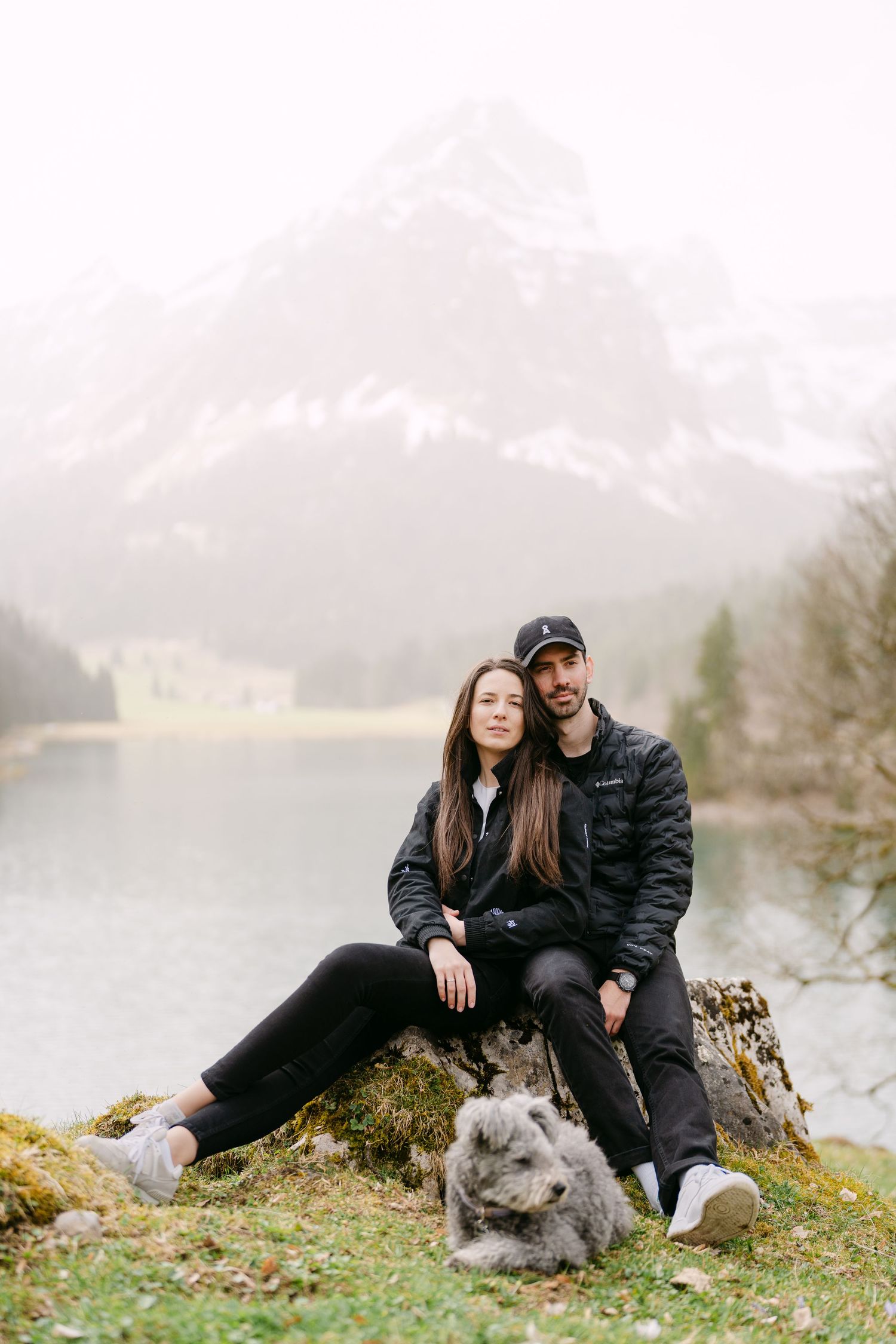 A couple sits with their grey dog on mossy rocks by a misty mountain lake.