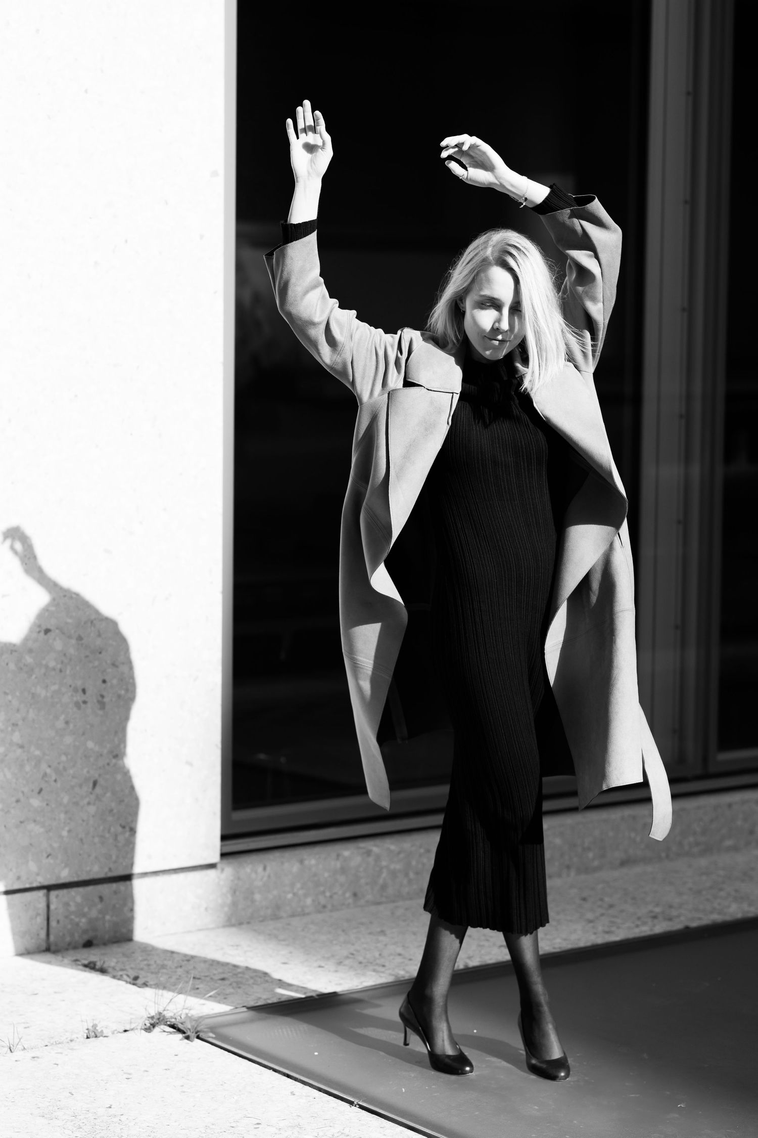 Black and white photo of woman in flowing coat striking dynamic pose against white wall.