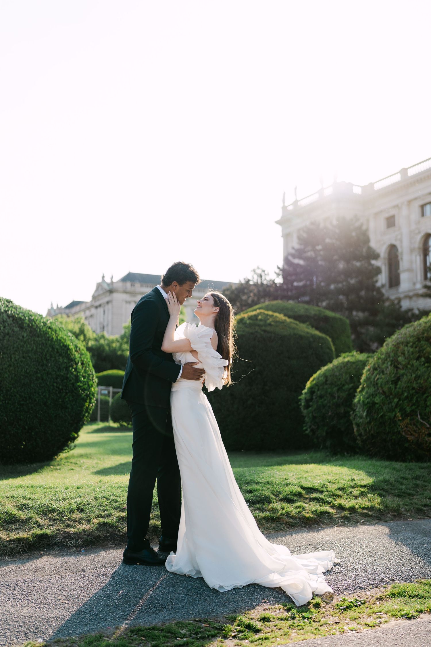Romantic wedding couple embraces in a garden with manicured hedges during golden hour sunset.