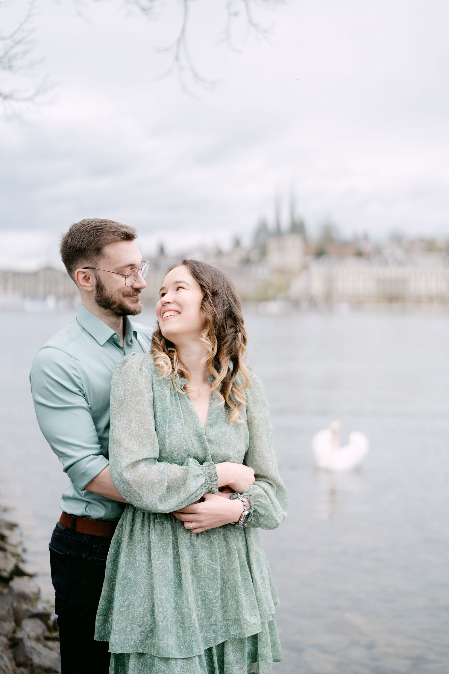 A couple in coordinating mint green outfits embrace by a waterfront with mountains in the distance.