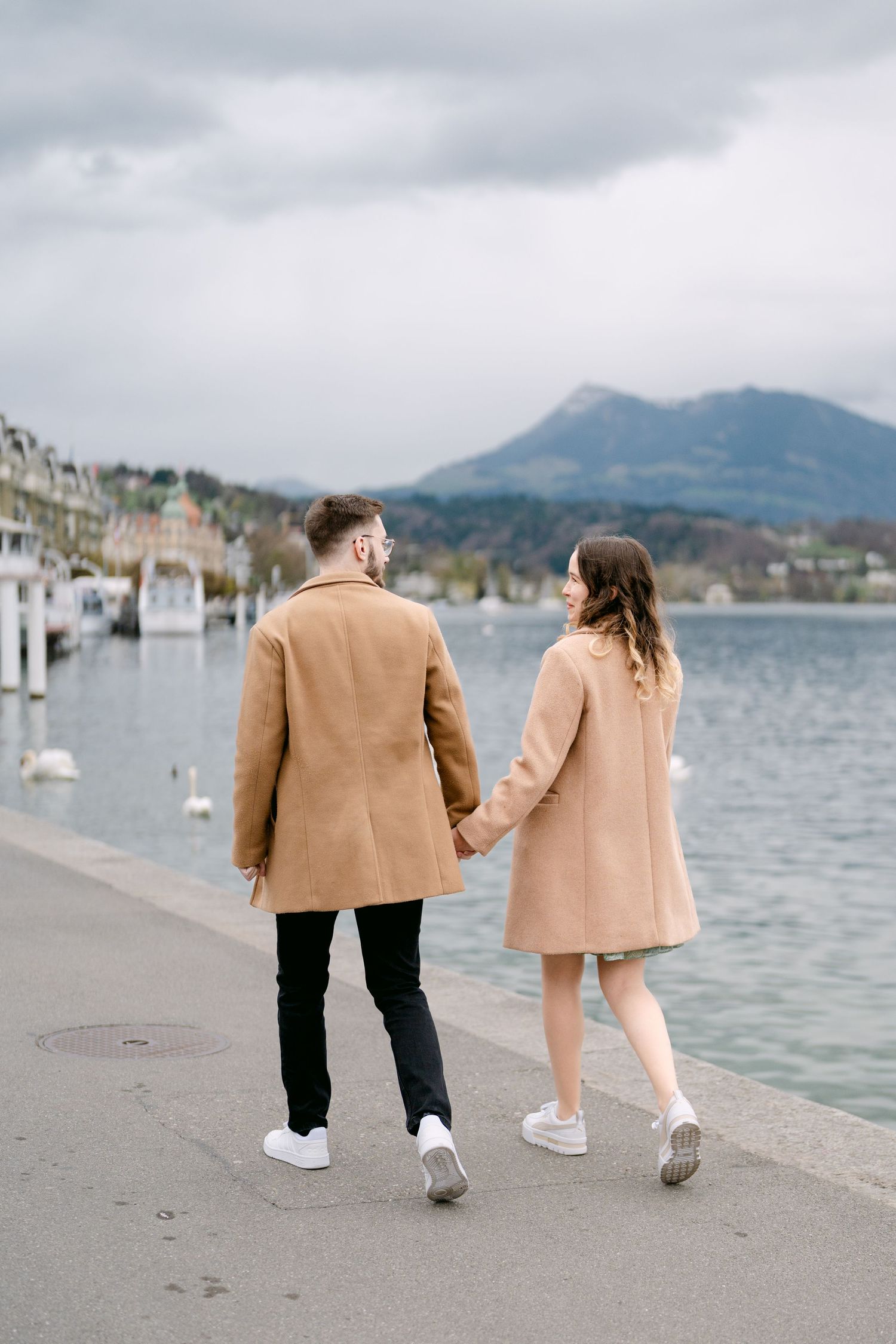 A couple in matching camel coats walks along a waterfront path with mountains and boats in view.