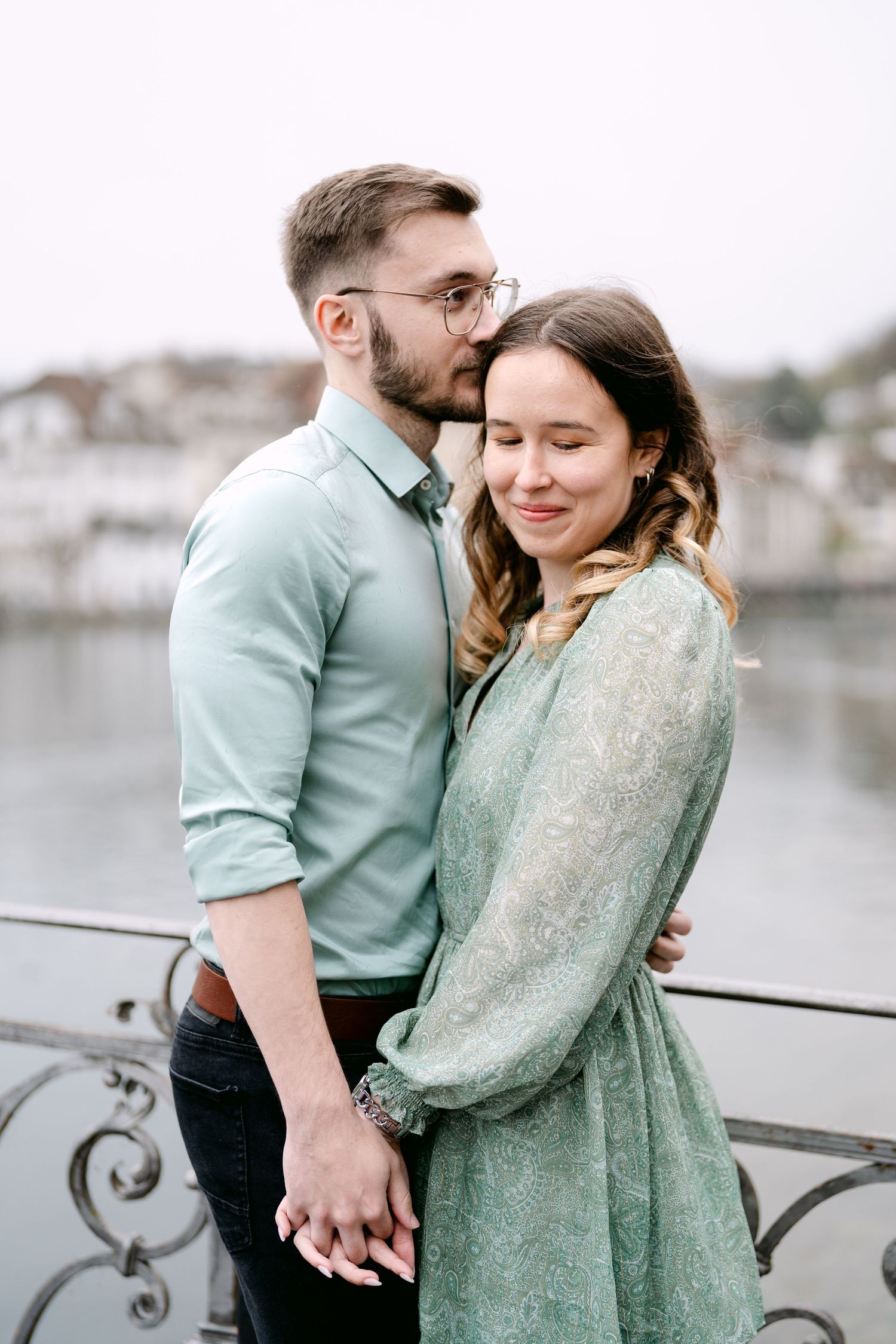 A romantic couple in matching mint green outfits embrace by a waterfront on a cloudy day.