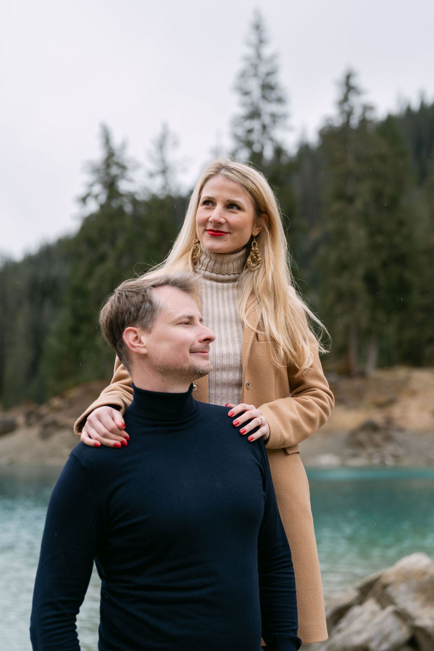 A couple poses by a turquoise alpine lake with evergreen trees in the background.