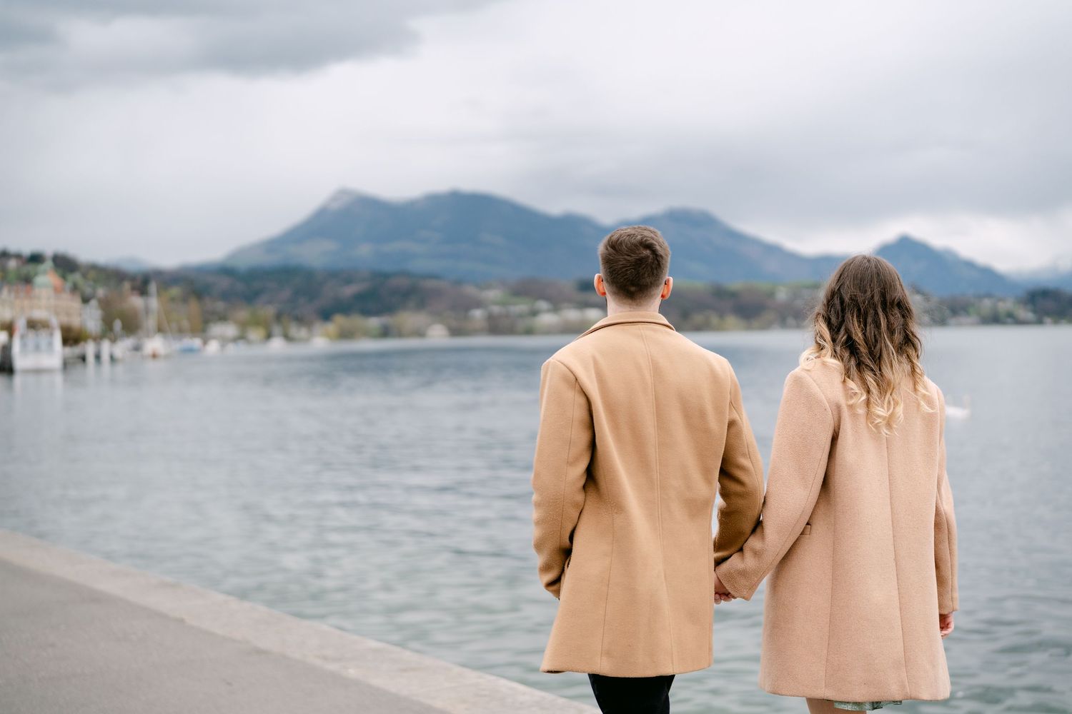 Couple in tan coats walking along a scenic waterfront with mountains in the background.