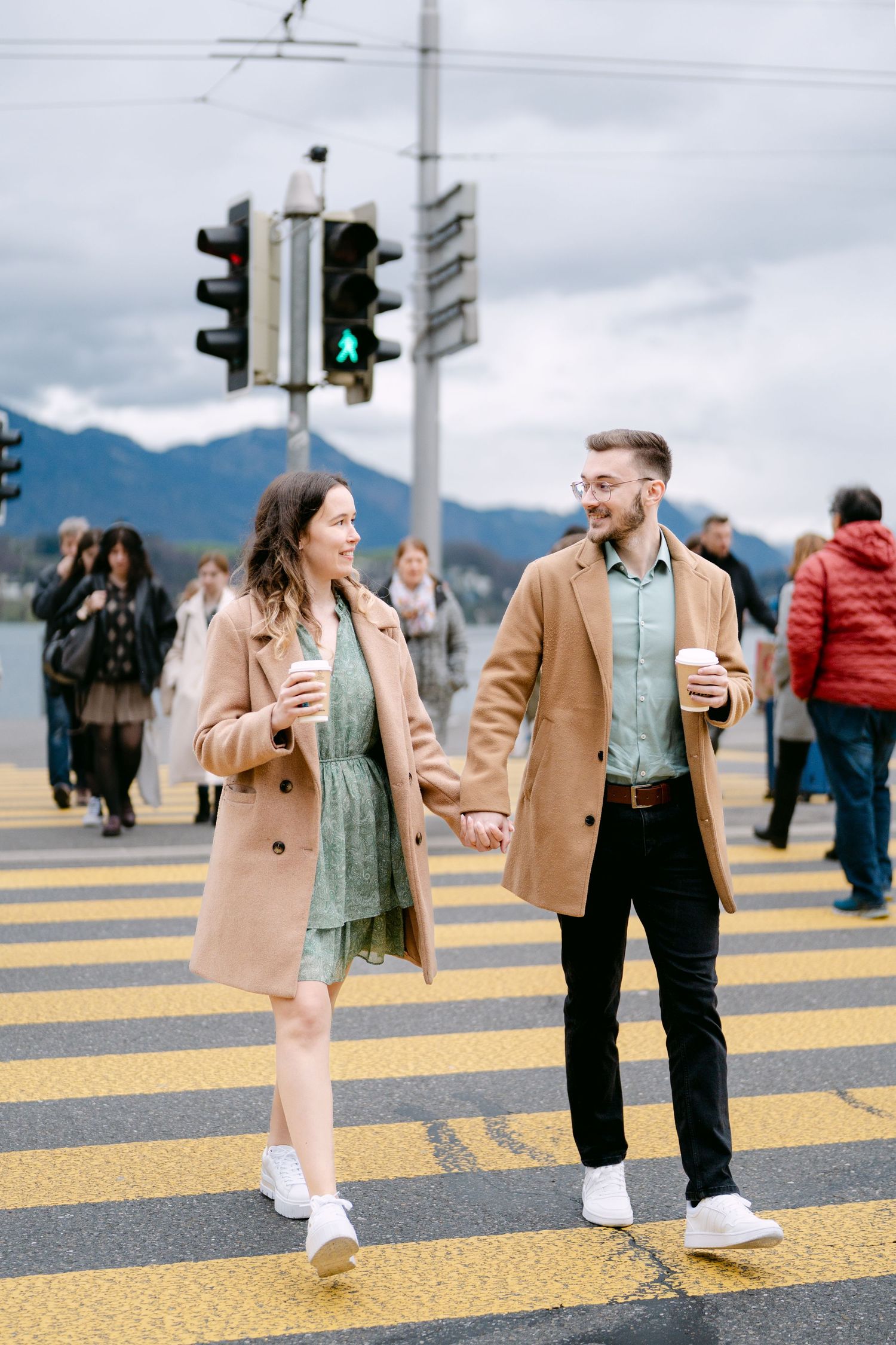 A couple in matching camel coats crosses a yellow pedestrian crossing with mountains in background.