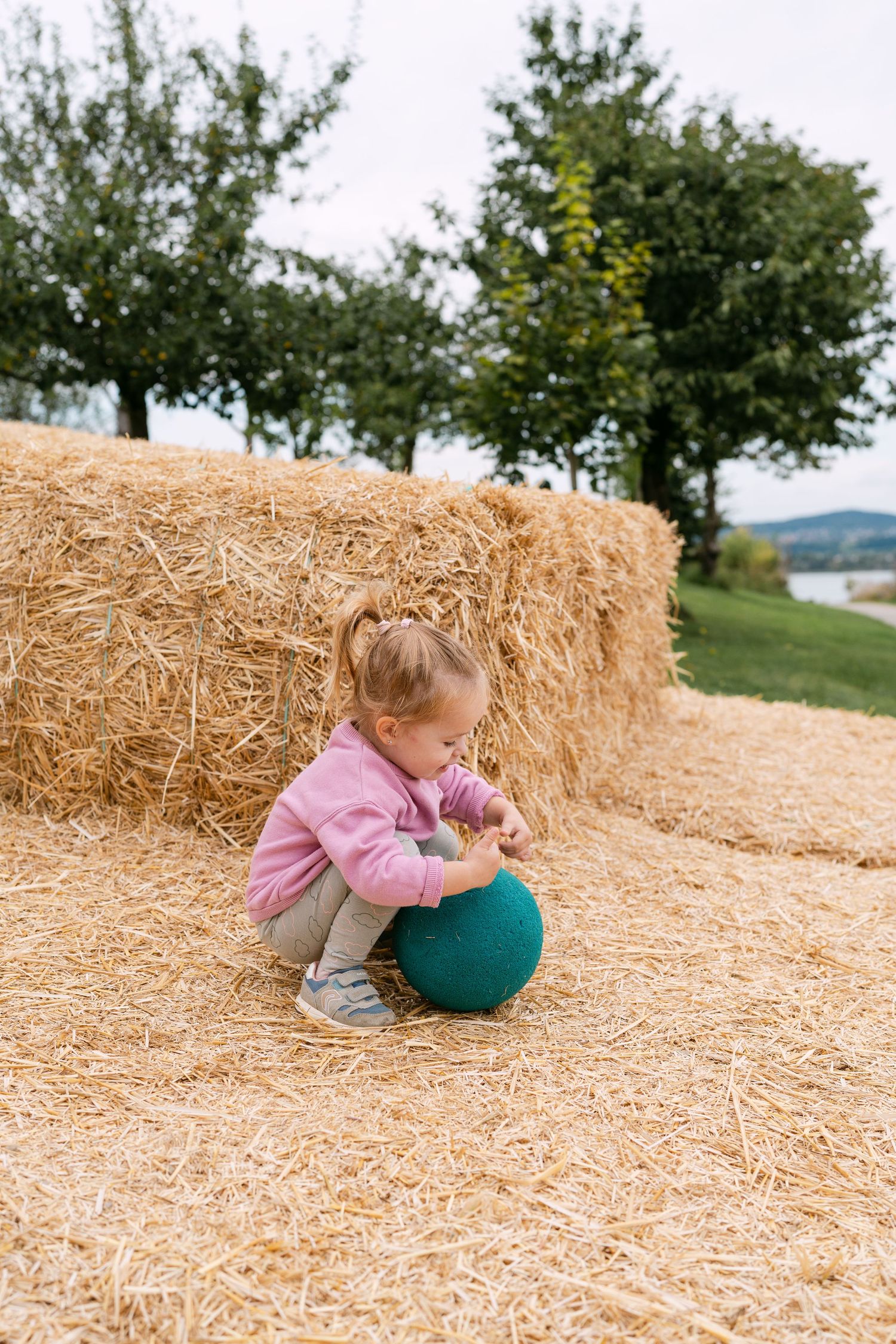 Child playing with a green ball on hay bales in an outdoor setting.